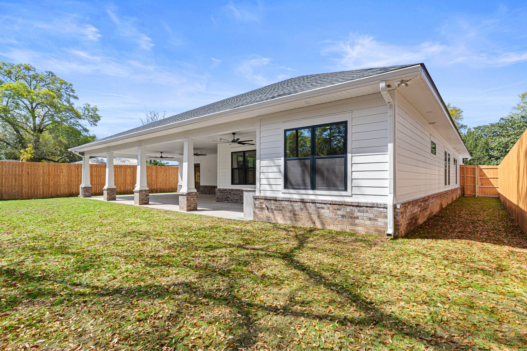 A modern house with white vinyl siding, a gray shingle roof, large black-framed windows, and a covered patio with ceiling fans, surrounded by a wooden fence and a grassy yard with fallen leaves.