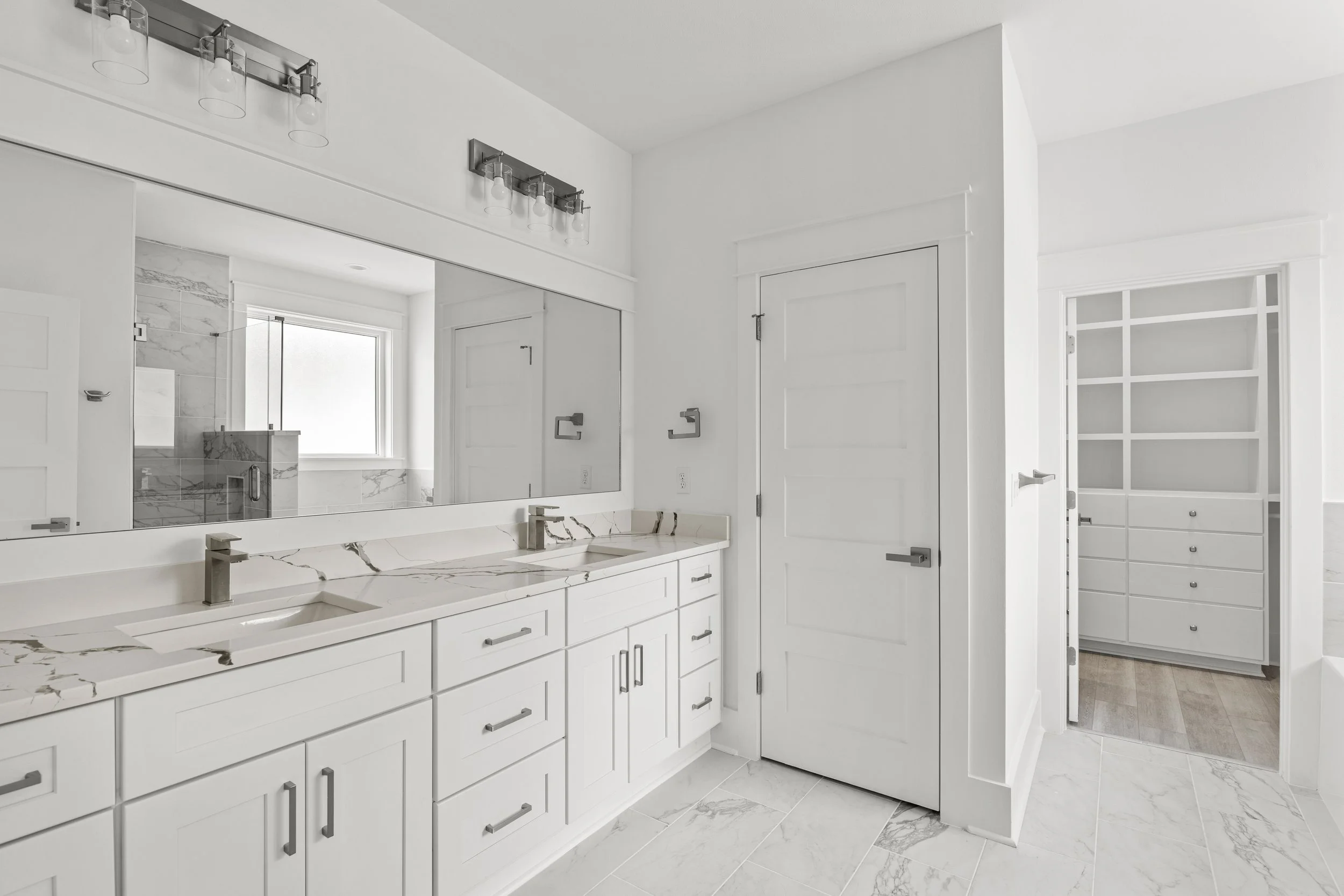 Modern white bathroom with marble countertops, double sink, large mirror, cabinets, and a walk-in shower with glass door.