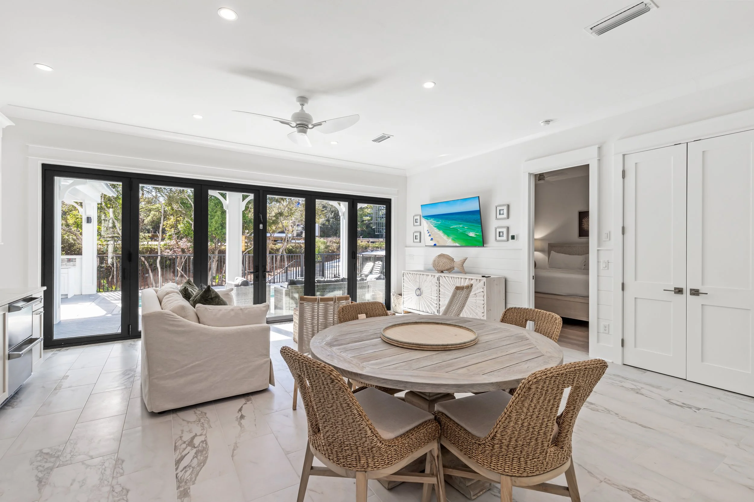 Living room with sliding glass doors opening to a balcony, white walls, ceiling fan, wall-mounted TV, and a round wooden dining table with wicker chairs.