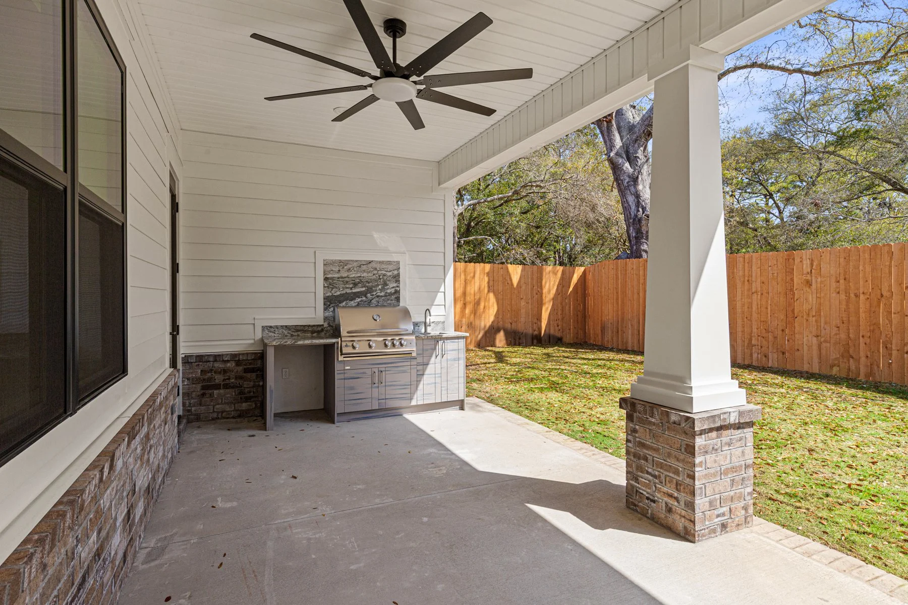 Covered outdoor patio with ceiling fan, built-in grill, and brick-built support columns. Fence and trees visible in the background.