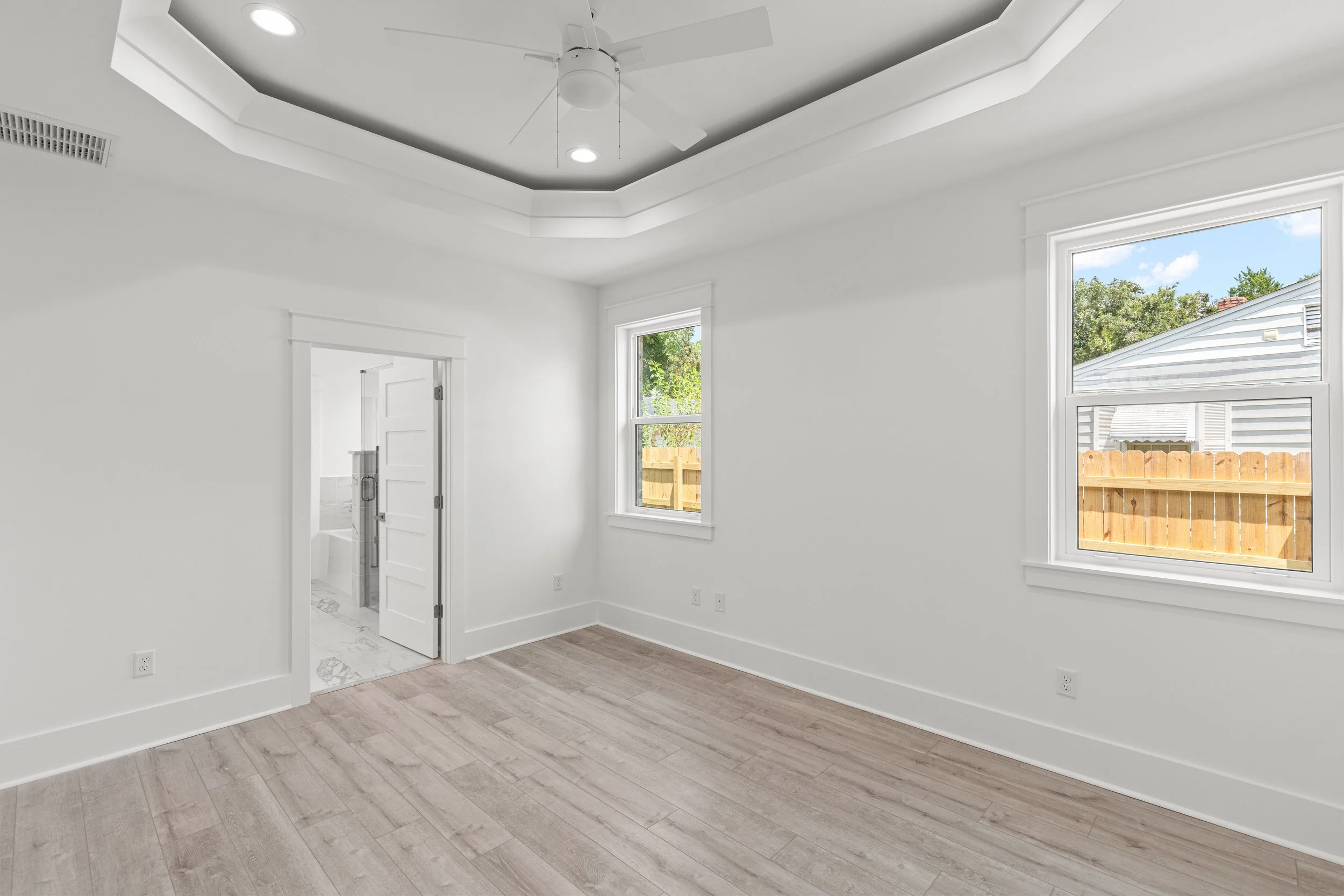 Empty room with white walls, wood flooring, two windows showing a fenced backyard, ceiling fan, and a door leading into another room.