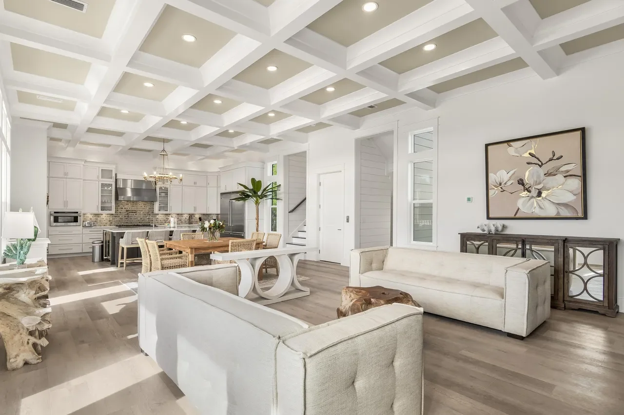 Bright and spacious open-concept living area with white sofas, wooden flooring, a dining table, and a modern kitchen with white cabinetry and stainless steel appliances.