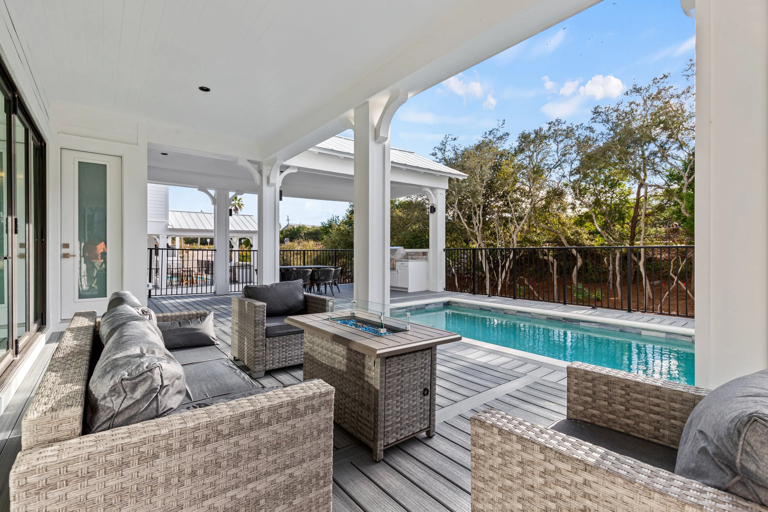 Outdoor patio area with wicker furniture, a small swimming pool, and a barbecue, overlooking trees and a partly cloudy sky.
