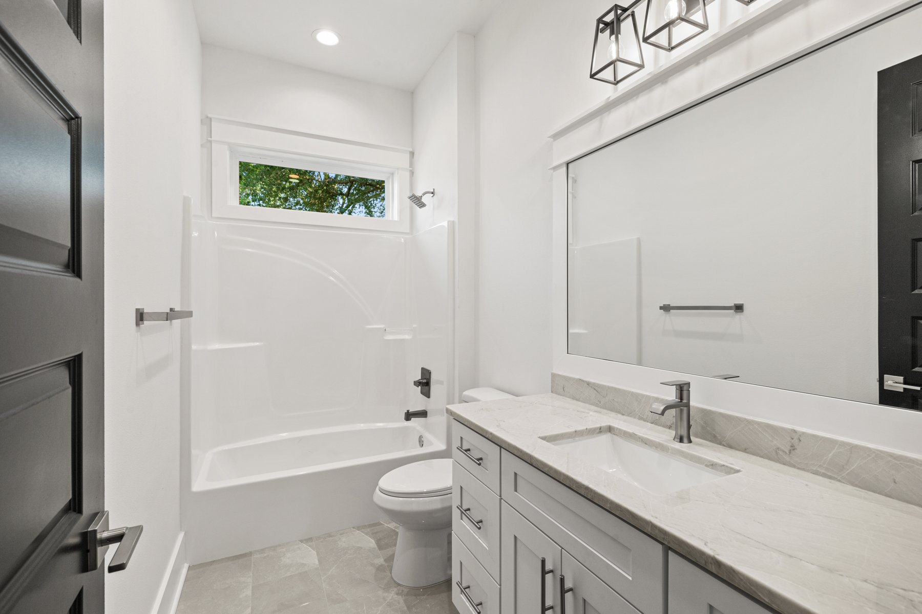 Modern bathroom with white walls, a bathtub with shower, a small horizontal window, a white vanity with a marble countertop and a rectangular sink, a large mirror, a black door, and [two pendant lights].