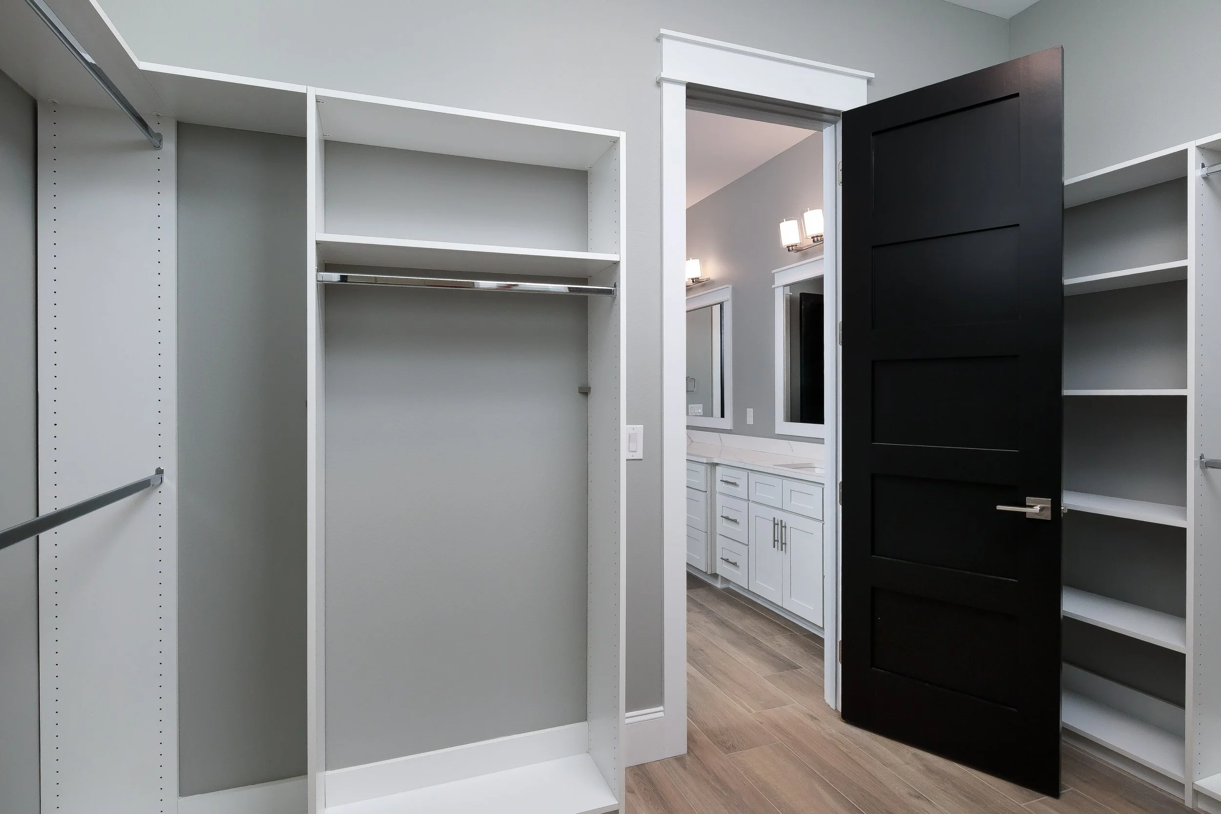 Empty white custom closet with metal hanging rods, open door leading to a bathroom with white cabinets, vanity lights, and a mirror.