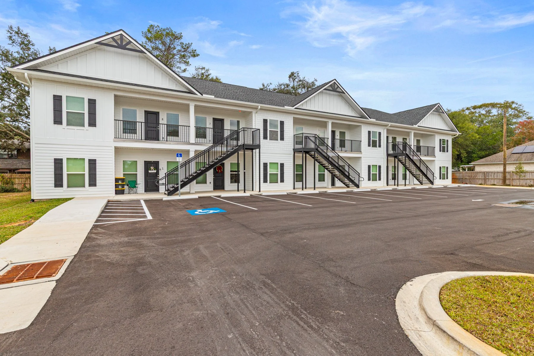 Exterior view of a modern two-story apartment complex with white siding, black shutters, and black staircases leading to the upper floor, surrounded by a parking lot with designated spaces including a handicapped spot, and a grassy area with trees in the background.