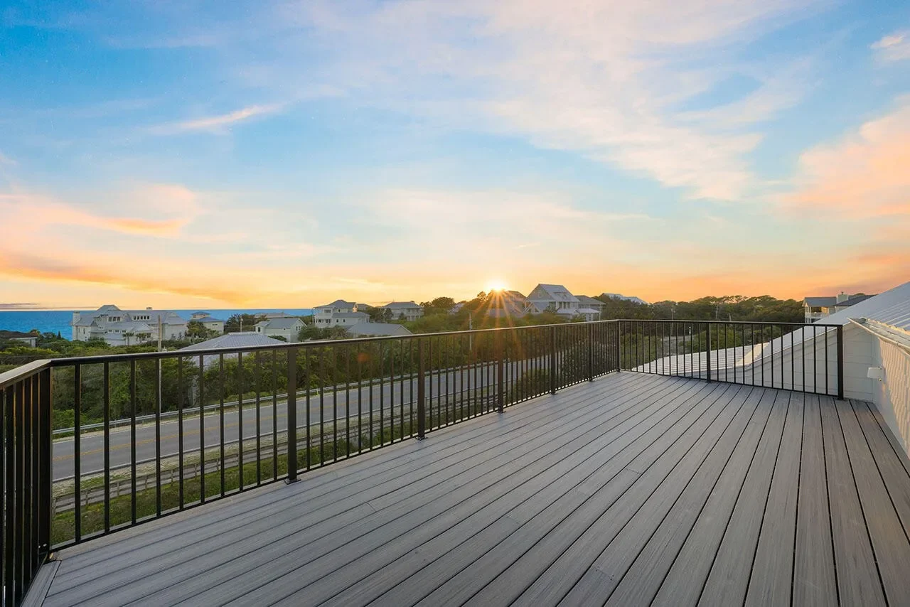 A spacious deck with a black railing overlooking a neighborhood with white houses and greenery during sunset.