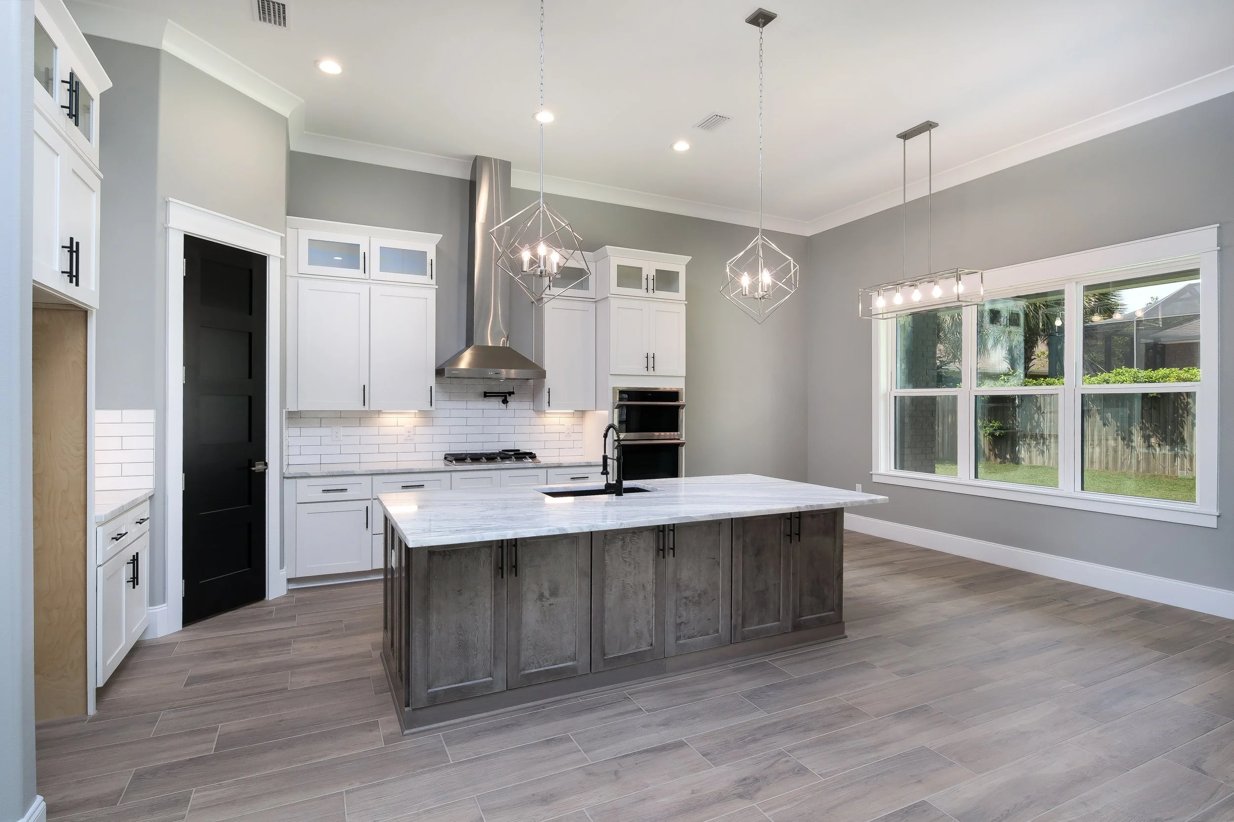 Modern kitchen with white cabinets, black handles, a gray island with a marble countertop, large window, and geometric pendant lights.