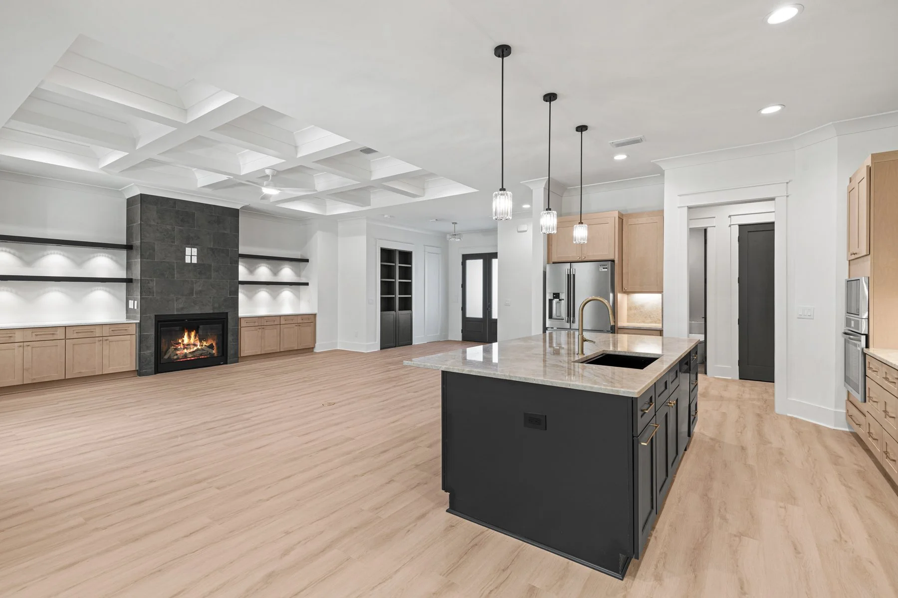 Modern open-plan kitchen and living area with hardwood floors, a black kitchen island, pendant lights, a fireplace with built-in shelving, stainless steel appliances, and a white coffered ceiling.
