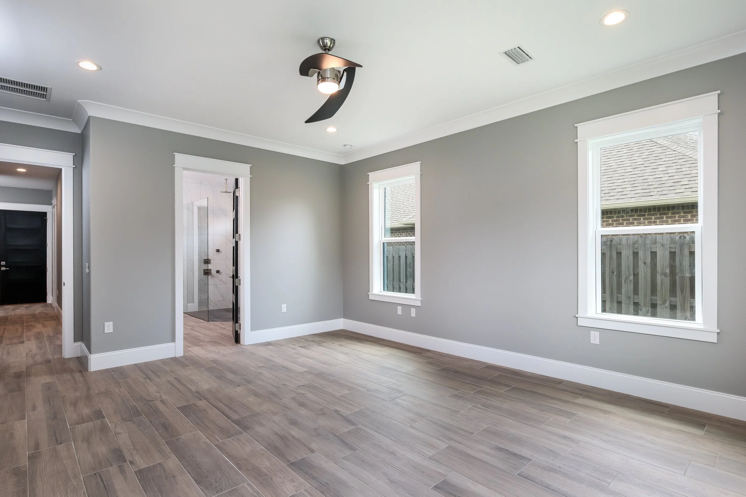 Empty modern living room with light-colored walls, large windows, wood-look flooring, and a ceiling light fixture.