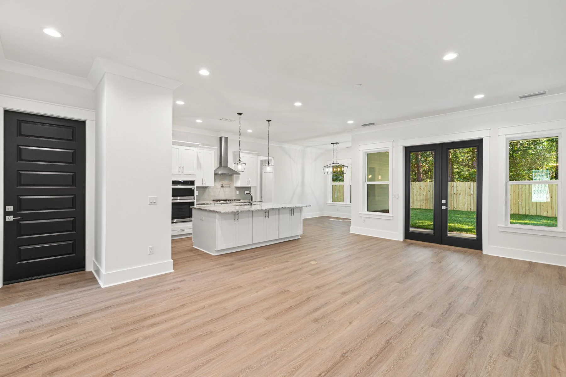 Open-concept kitchen and living area with white cabinets, granite countertops, black front door, large windows, and wooden floor.