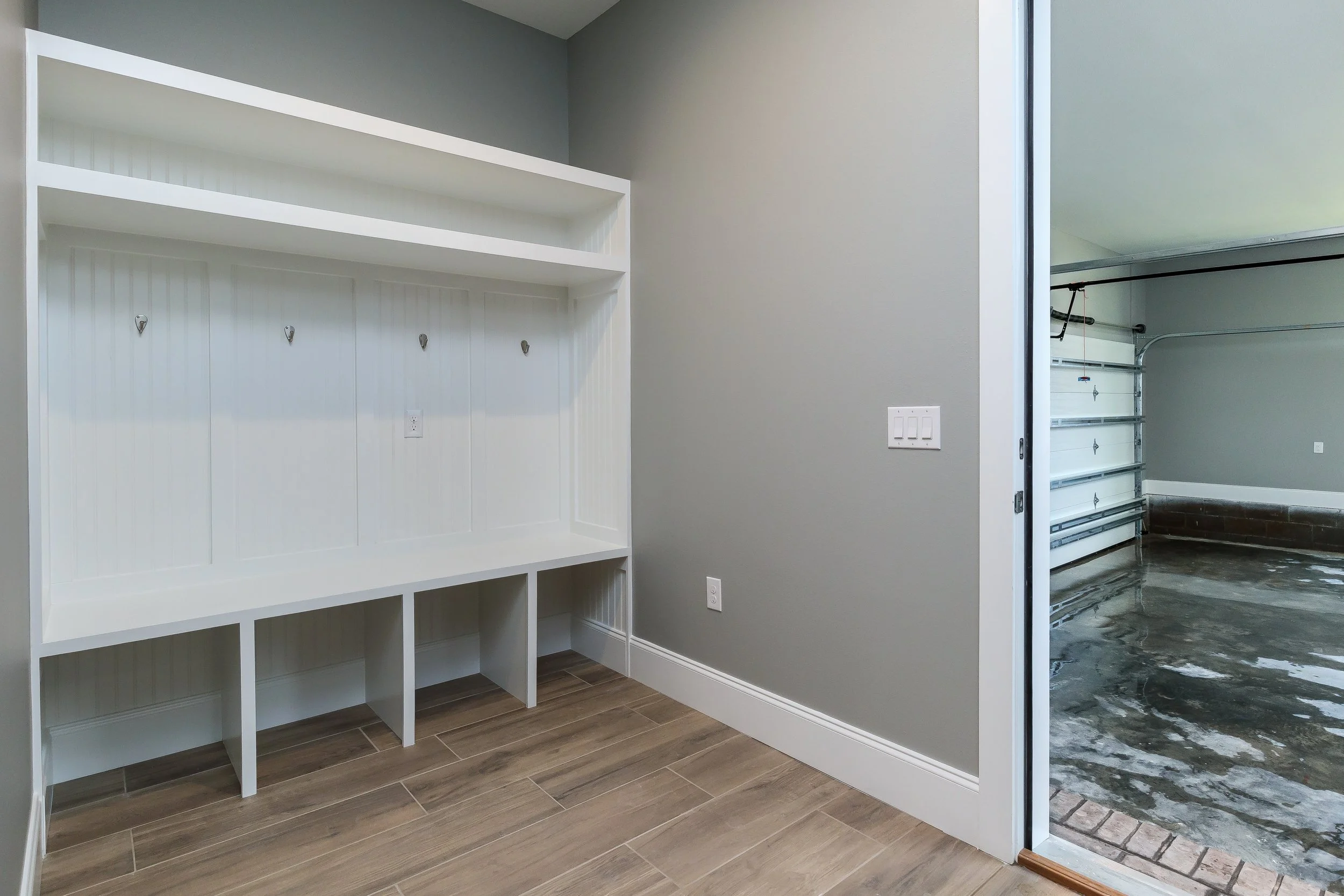 Custom built-in white mudroom shelf with hooks, a gray wall, and a doorway leading to a garage with a concrete floor.