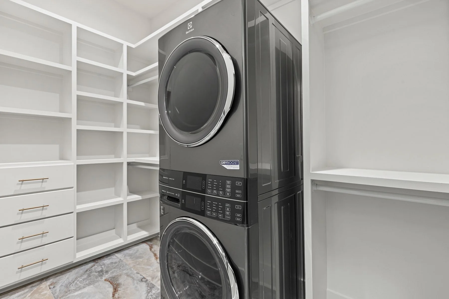 A laundry room with white shelving and drawers on the left, and a dark gray Electrolux front-loading washer and dryer stacked vertically on the right, with a marble floor.