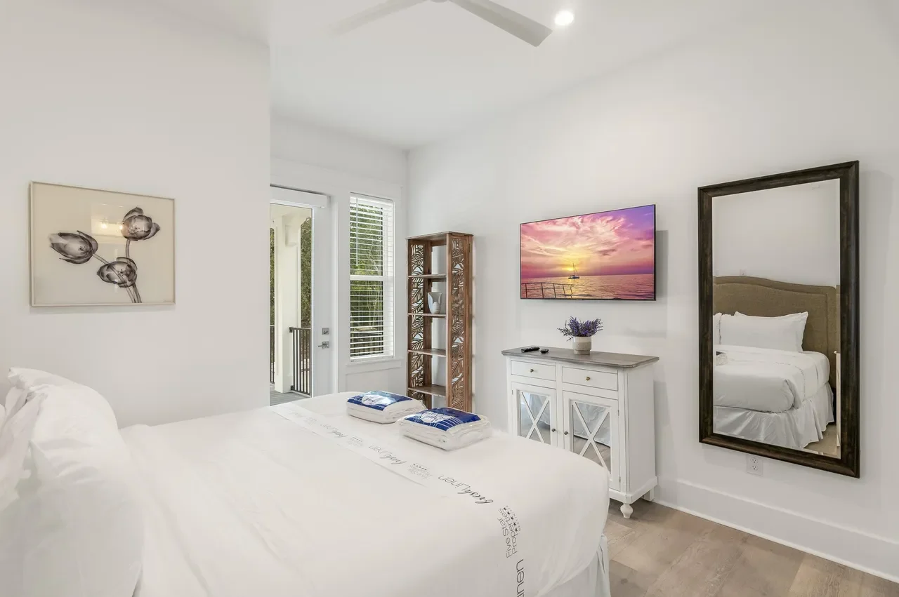 Bright hotel room with white walls, a large bed with white linens, a wall-mounted flat-screen TV showing a sunset, a mirror reflecting the bed, a wooden shelving unit, and outdoor balcony doors with white blinds.