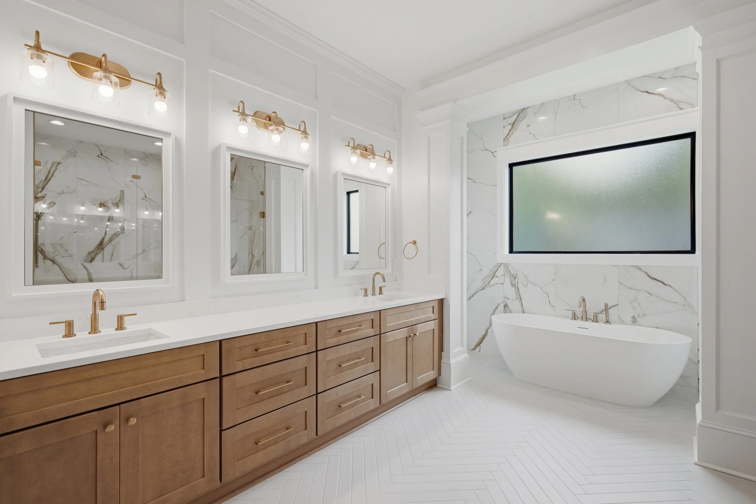 Modern bathroom with white marble walls, large frosted window, wooden vanity with gold fixtures, and a freestanding bathtub.