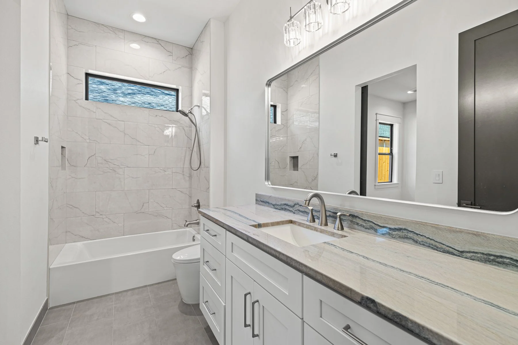 Modern bathroom with white vanity, marble countertop, large mirror, gray tiled floors, marble shower with a small horizontal window, glass light fixtures, and a toilet.