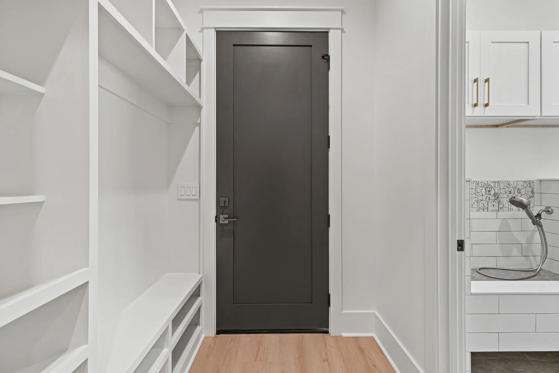 View of a narrow hallway with white built-in shelves on the left, a gray door in the center, and a glimpse of a bathroom with a small shower area on the right.