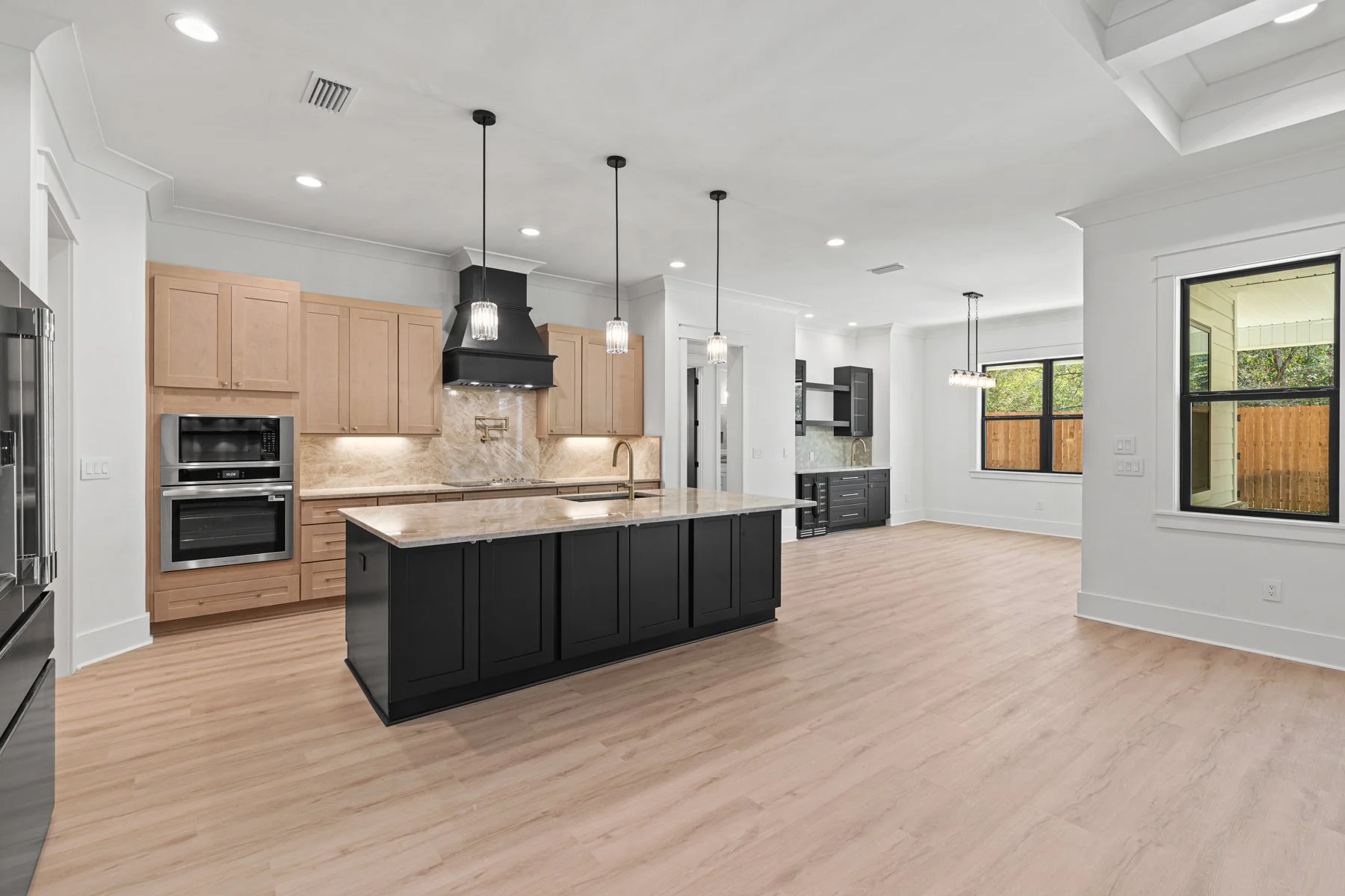 Modern kitchen with black and wooden cabinets, an island with a marble countertop, pendant lighting, large windows showing a backyard, hardwood floors, and a partially visible outside patio.