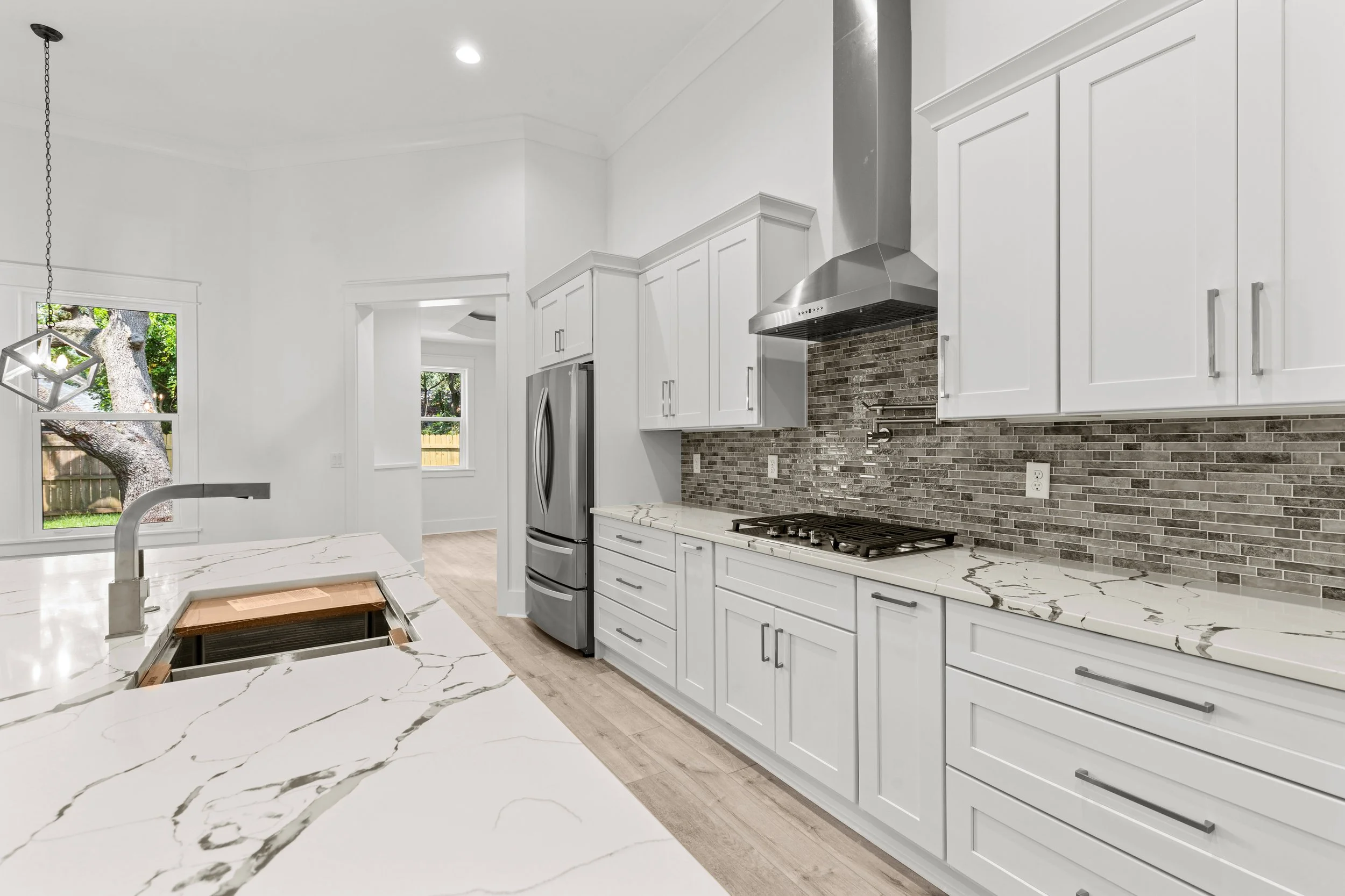 Modern kitchen with white cabinets, marble countertop, brick backsplash, stainless steel range hood, and refrigerator, with windows showing outdoor trees.
