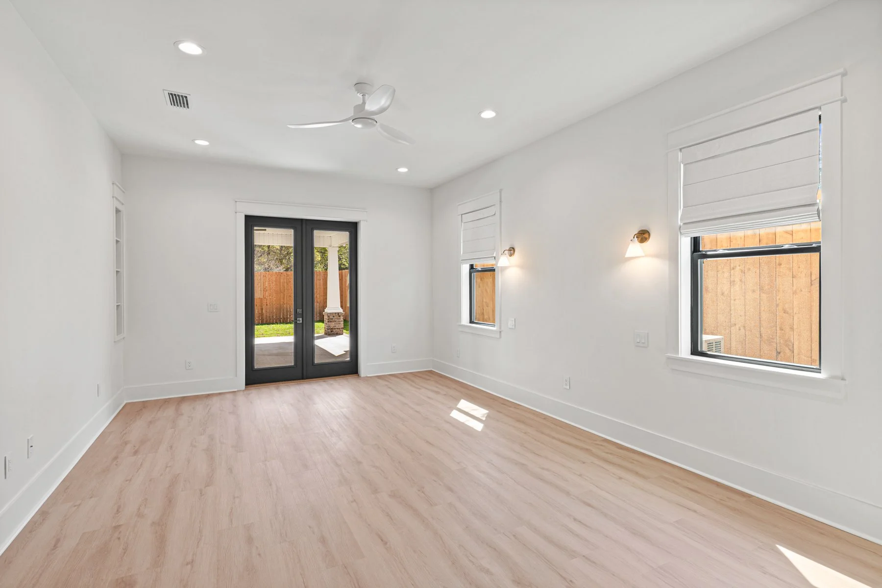 Empty living room with light wood flooring, white walls, two windows with white shades, a ceiling fan, recessed lighting, and a glass door leading to a backyard with grass and a wooden fence.