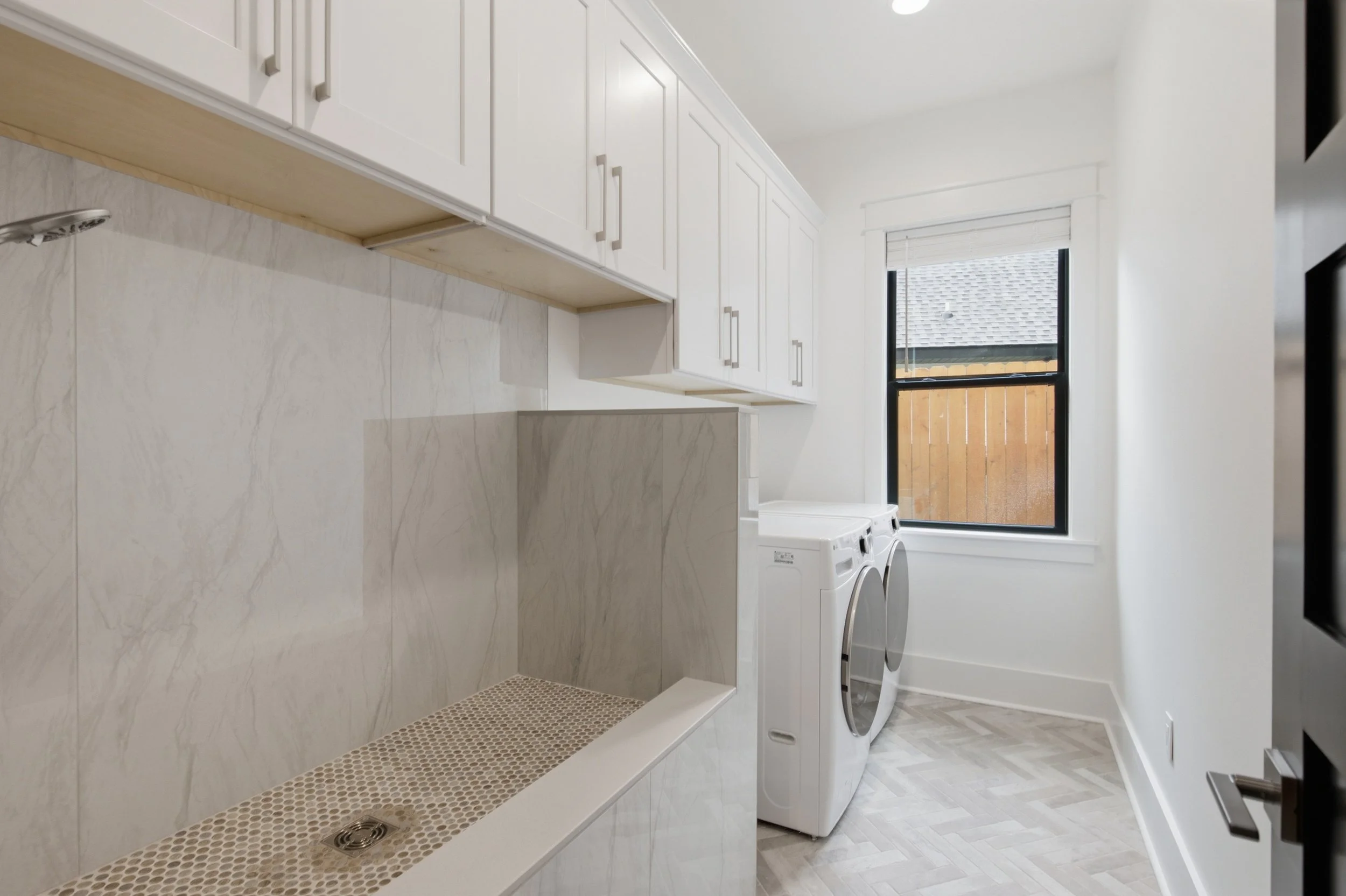 A laundry room with white cabinets, a metal vent in the wall, a washer and dryer, a window with a blind, and a tiled floor.