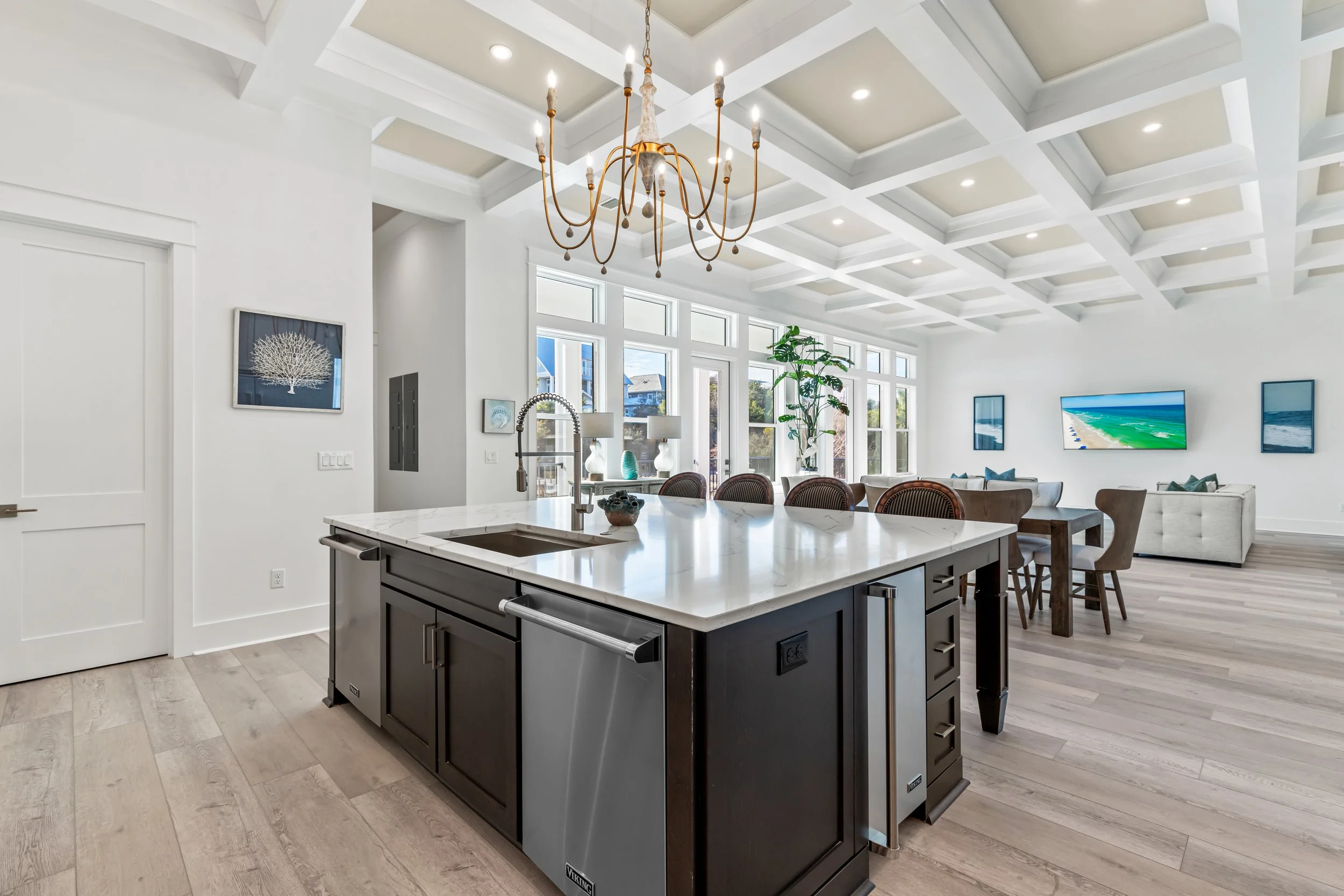 A spacious, modern open-concept kitchen and living area with white walls and large windows, featuring a central kitchen island with a marble countertop, dark cabinetry, and a stainless steel sink. The room has a high coffered ceiling with a large chandelier, and the living area has a wall-mounted TV, artwork, and seating in front of the windows.
