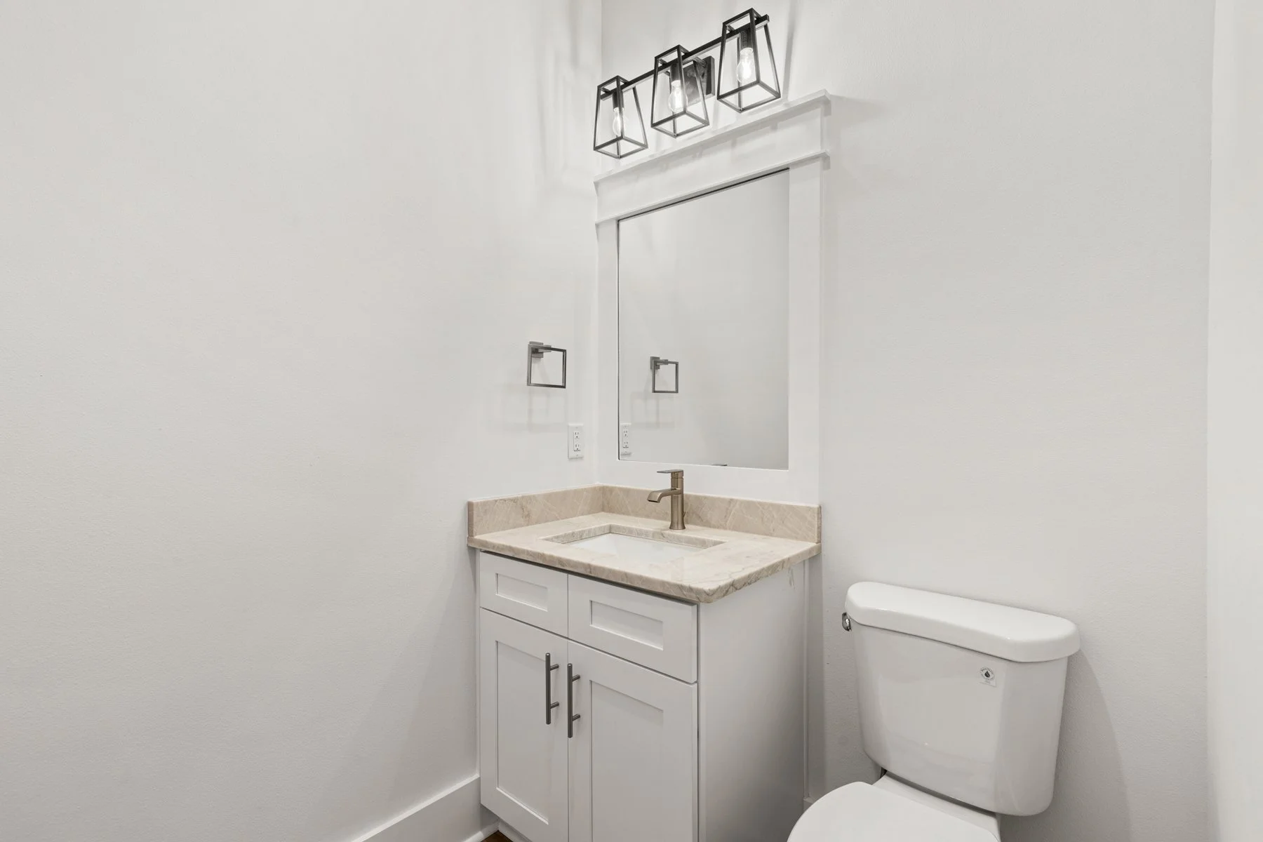 A small bathroom with white walls, a white vanity with a beige marble countertop, a rectangular mirror, black geometric light fixtures, a silver faucet, a white toilet, and simple black towel holders.
