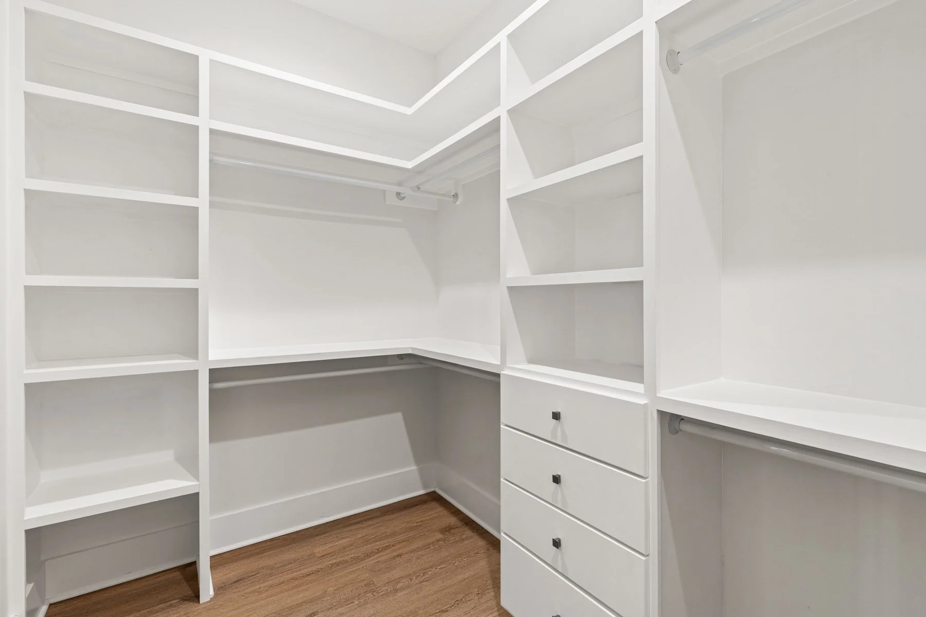 Empty walk-in closet with white built-in shelves and drawers, wood flooring.