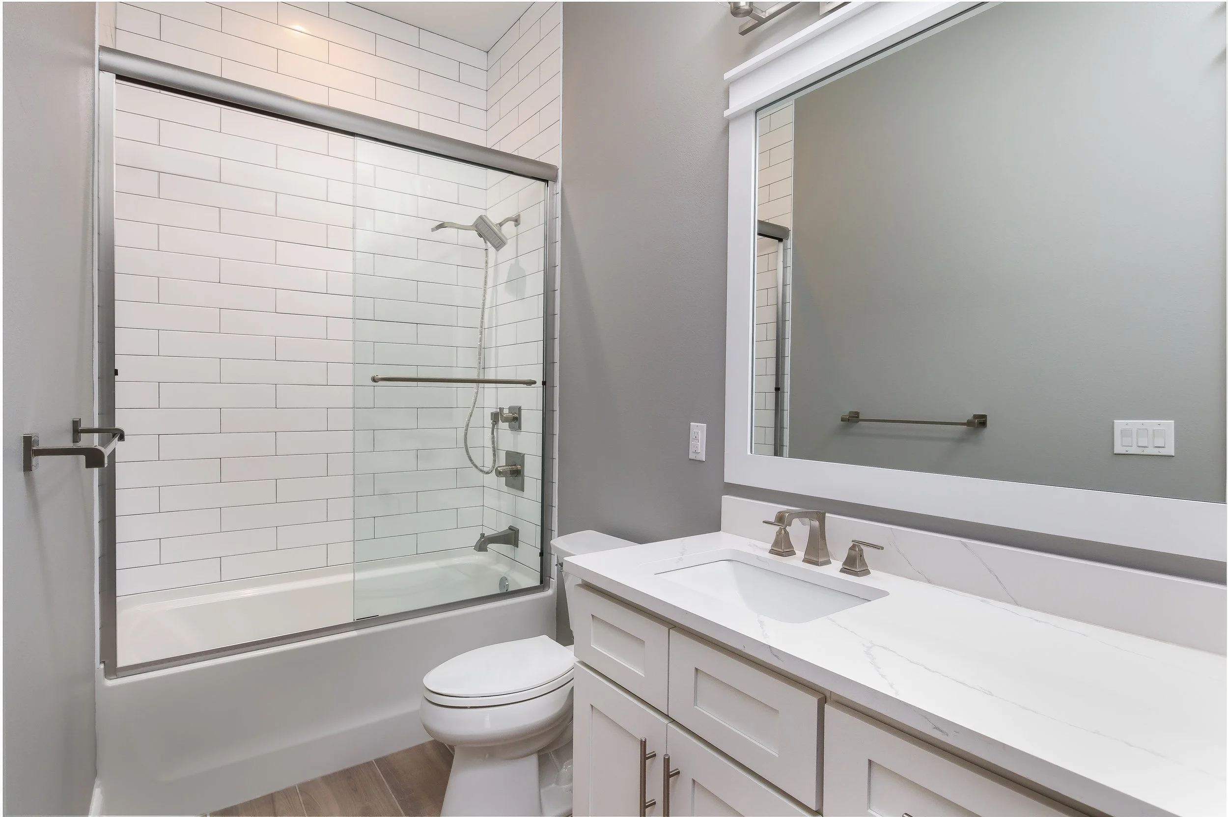 Modern bathroom with a glass-enclosed shower featuring white subway tiles, a gray wall, a large mirror, white vanity with a double sink, and a toilet.