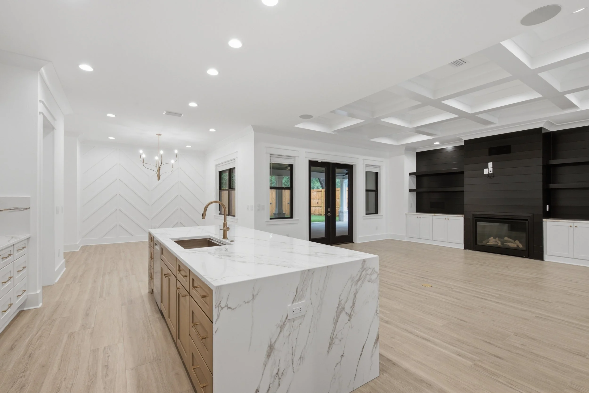 A modern kitchen with white walls and ceiling, light wood flooring, a marble island with tan cabinets, black sliding doors leading outside, a black fireplace with a mounted TV above, and a ceiling with square tray details and recessed lighting.