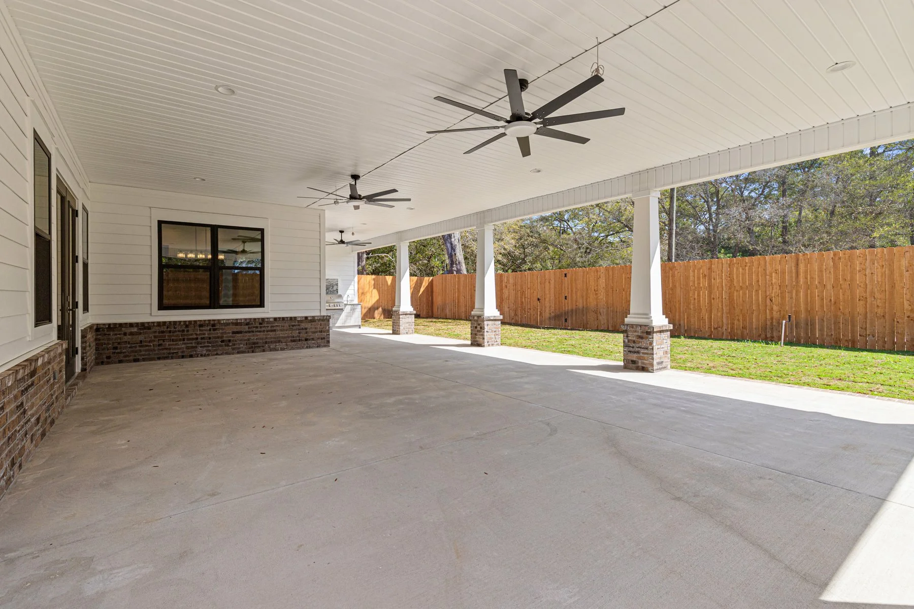 Covered patio with white ceiling, ceiling fans, windows, brick accents, concrete flooring, and a wooden fence surrounding a backyard.