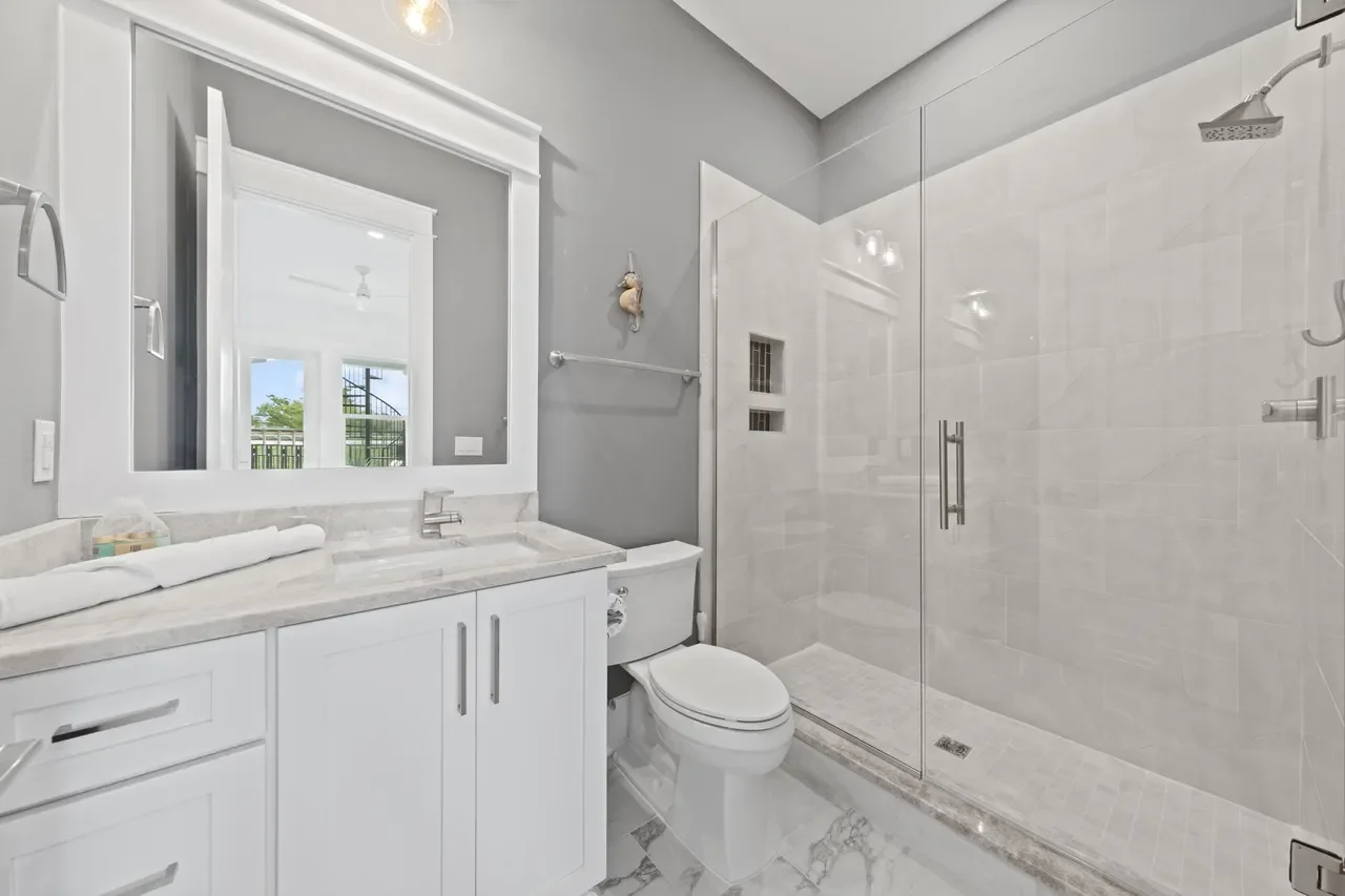 Modern bathroom with a glass-enclosed shower, white vanity with a marble countertop, a mirror, and a toilet. The wall is painted gray, and there's a window with a view outside.