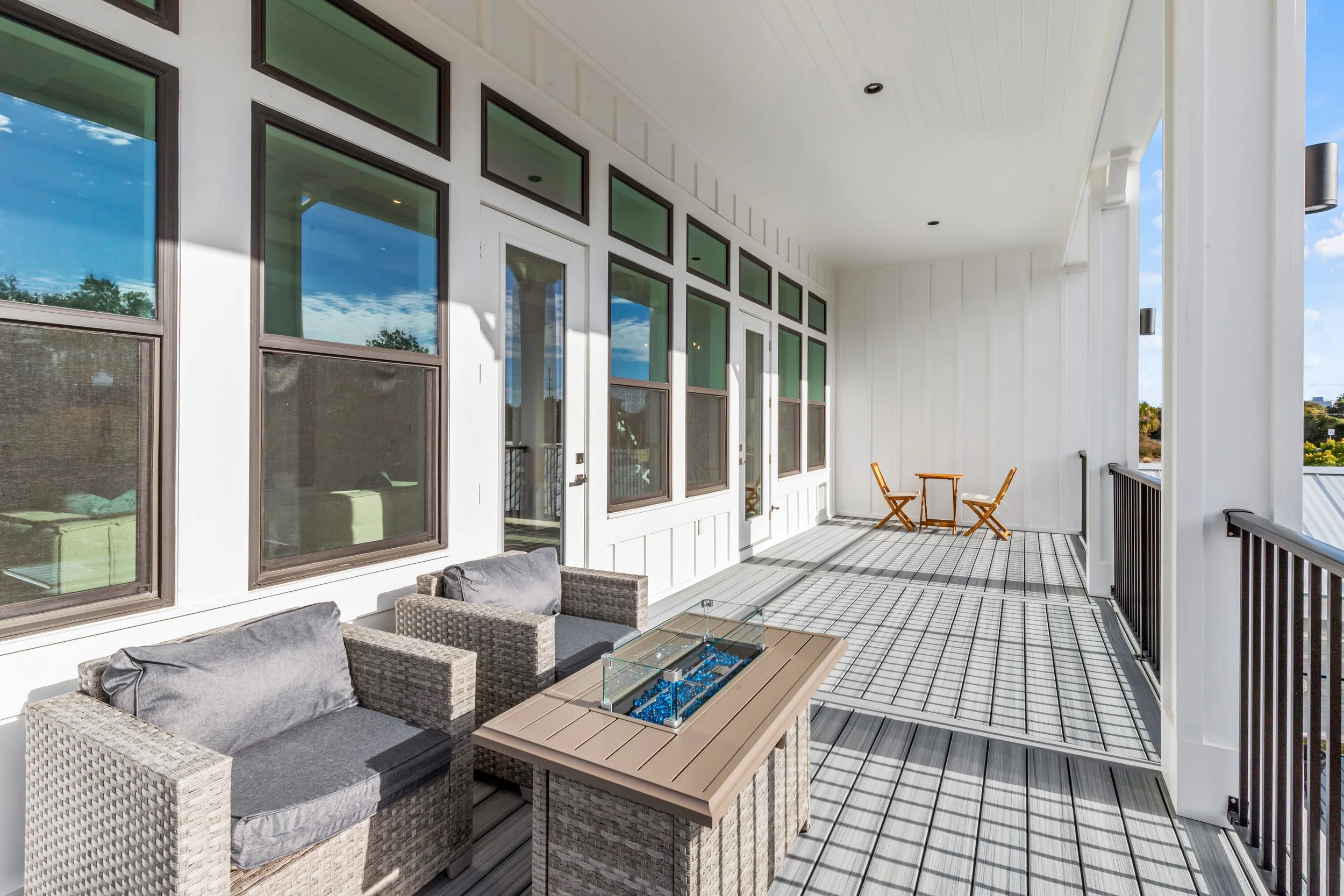 Balcony with outdoor seating, including wicker chairs with cushions and a table with a glass cover, overlooking a blue sky with white clouds and green trees.