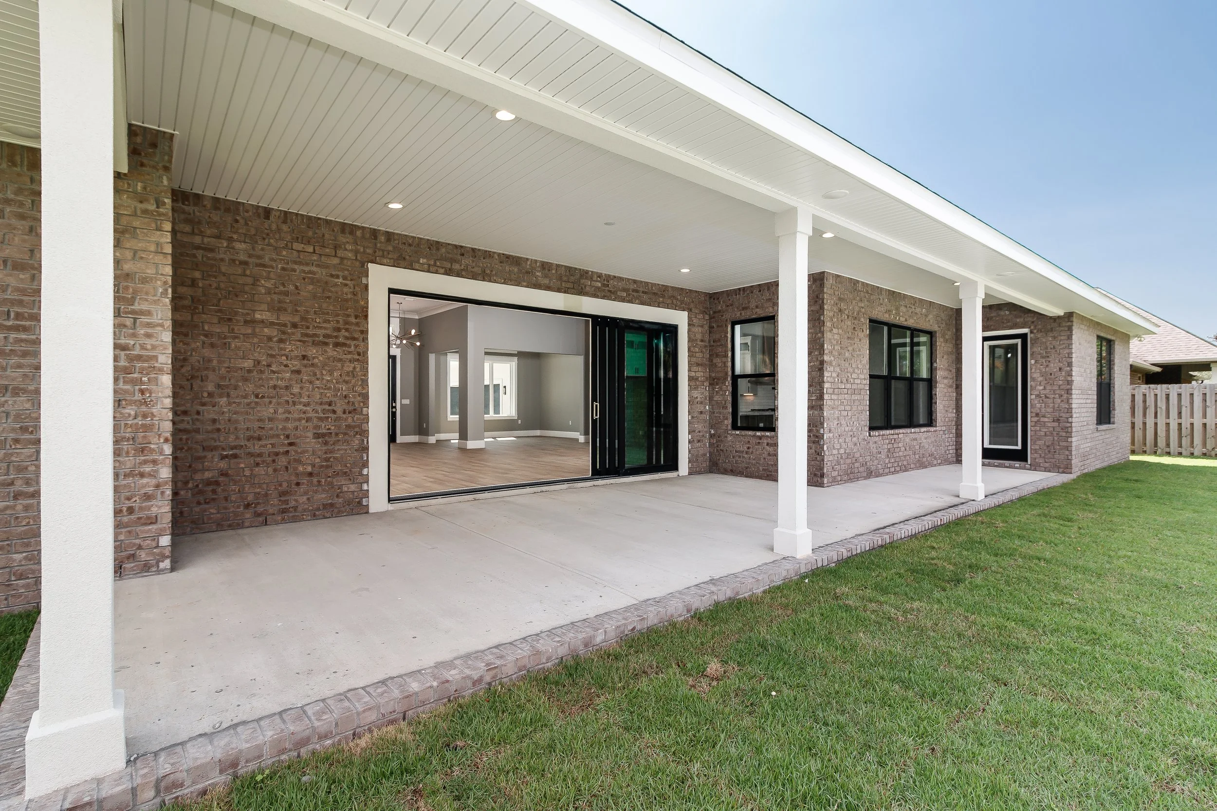 Backyard patio area with a covered porch, brick exterior, sliding glass door, and interior visible through the open door, with a lawn and neighboring houses in the background.