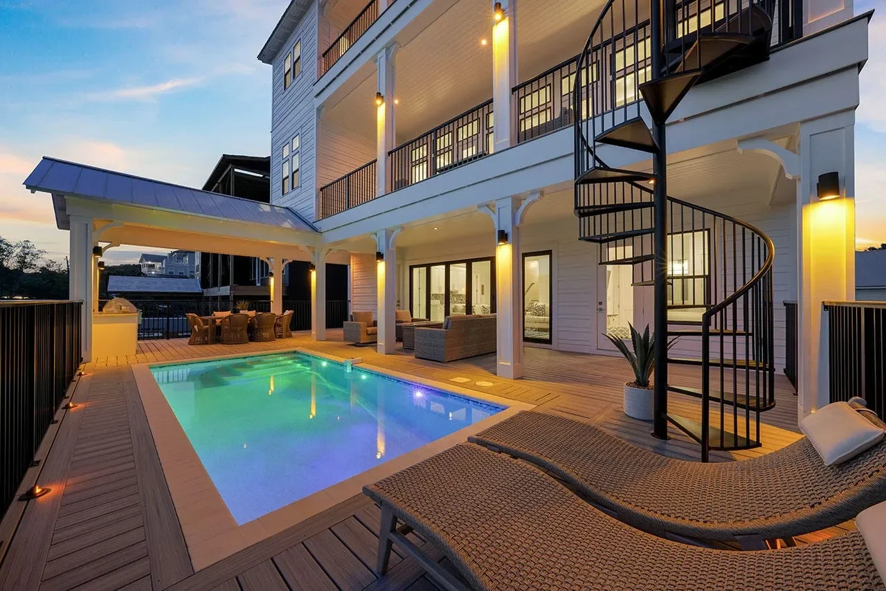 Outdoor backyard with a lit swimming pool, lounge chairs, patio furniture, spiral staircase, and hanging lights at sunset.