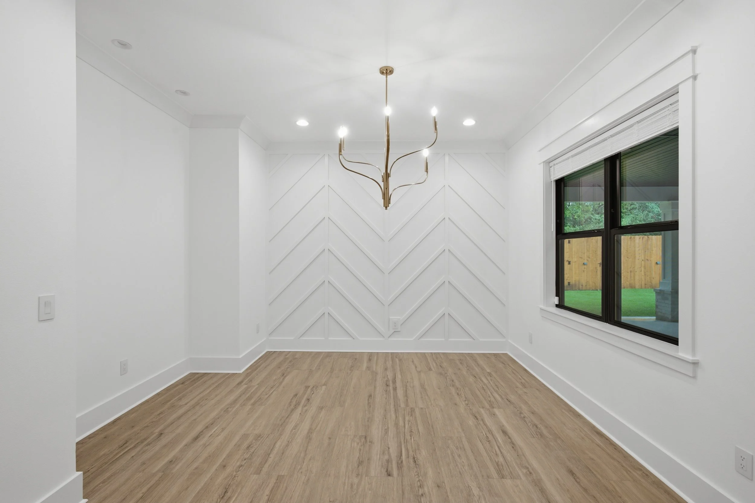 Empty room with white walls, wood flooring, a modern brass chandelier, and a window showing a backyard with a wooden fence and trees.