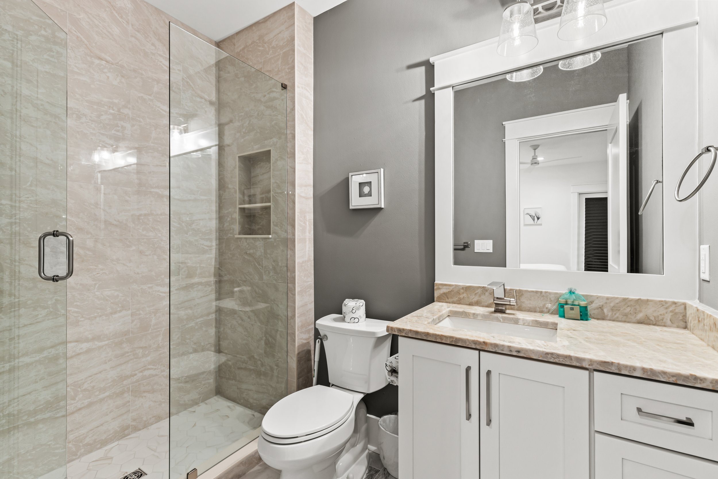 A modern bathroom with a glass-enclosed shower, a white toilet, and a white vanity with a marble countertop. The walls are painted gray, and a large mirror is above the sink. There are wall-mounted light fixtures above the mirror and a small framed picture on the wall. Items like soap and toiletries are on the countertop.