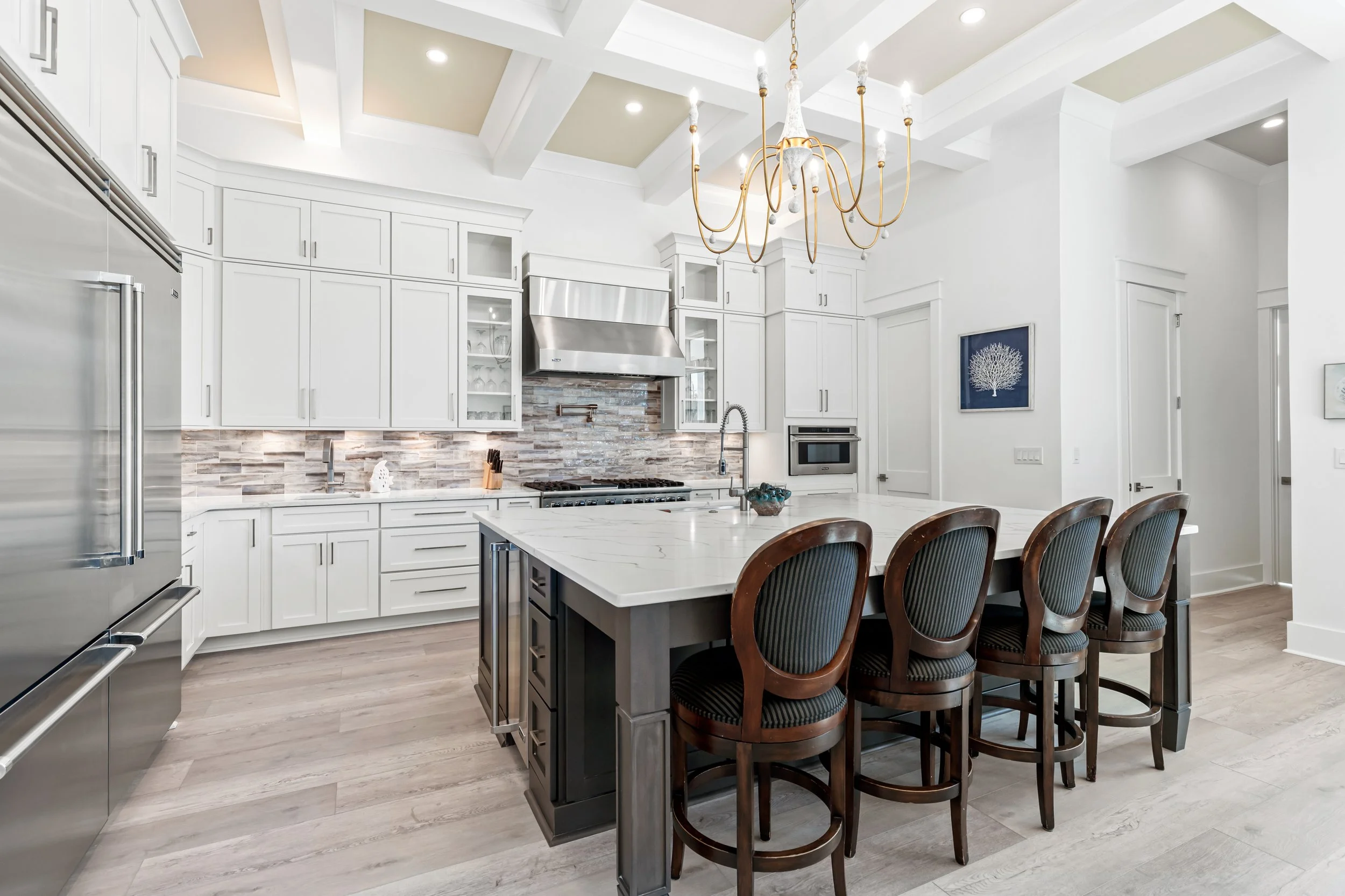 Modern kitchen with white cabinets, stainless steel appliances, and a white island with dark wooden chairs. A chandelier hangs from the ceiling, and there is artwork on the wall.