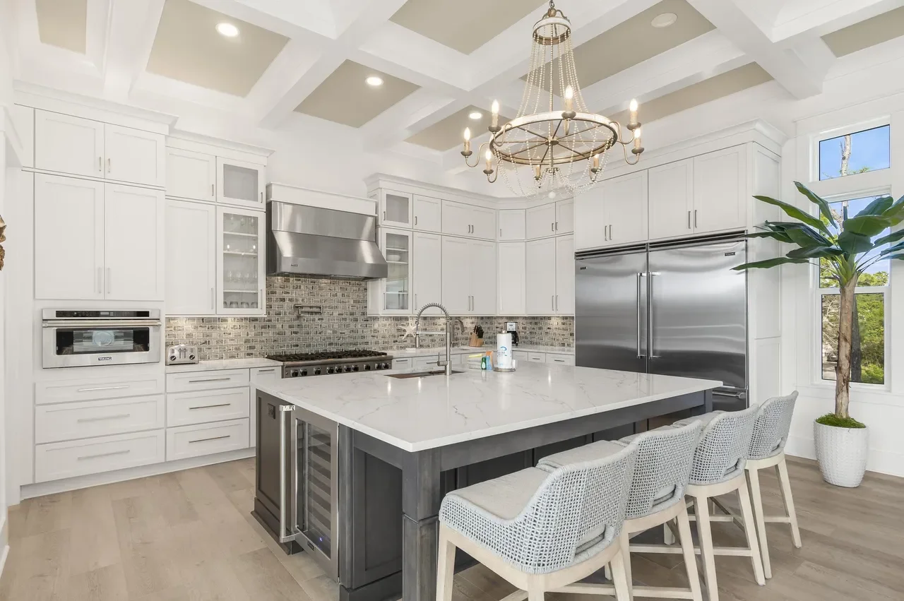 Modern white kitchen with marble island, stainless steel appliances, and a chandelier, featuring a potted plant by a window.