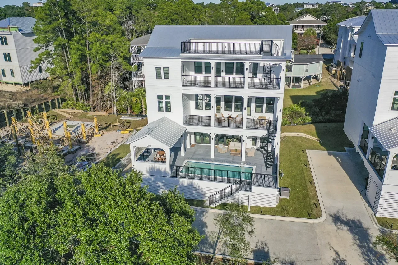 A multi-story white house with balconies, a swimming pool, and outdoor seating in a residential neighborhood with other similar houses and green trees.