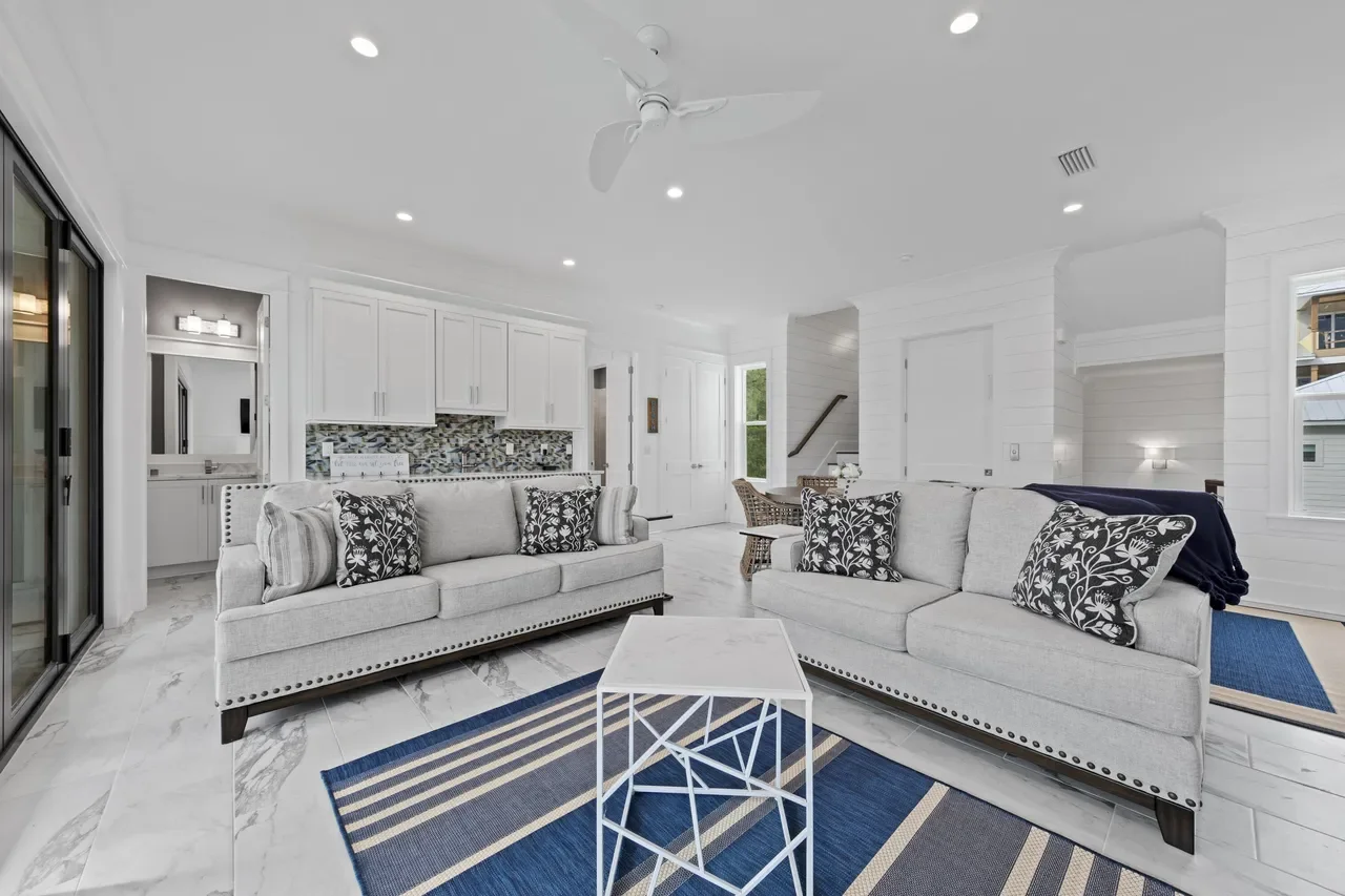 Bright living room with white walls, white ceiling, two light gray sofas with black and white patterned pillows, a white side table, a blue striped rug, a ceiling fan, and large windows letting in natural light.