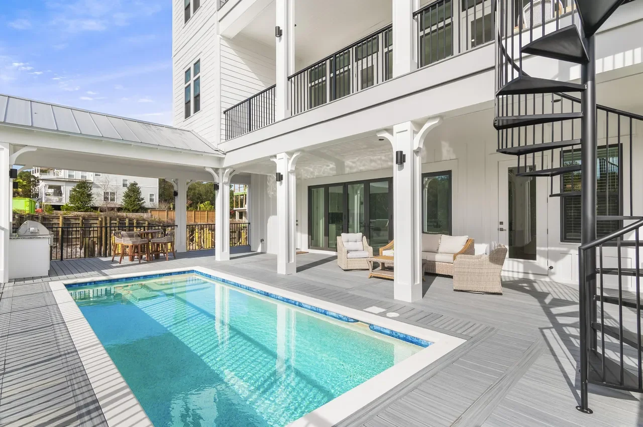 Backyard with a swimming pool, outdoor seating area with wicker chairs and a small table, sliding glass doors, white house exterior, open balcony, and a black spiral staircase.