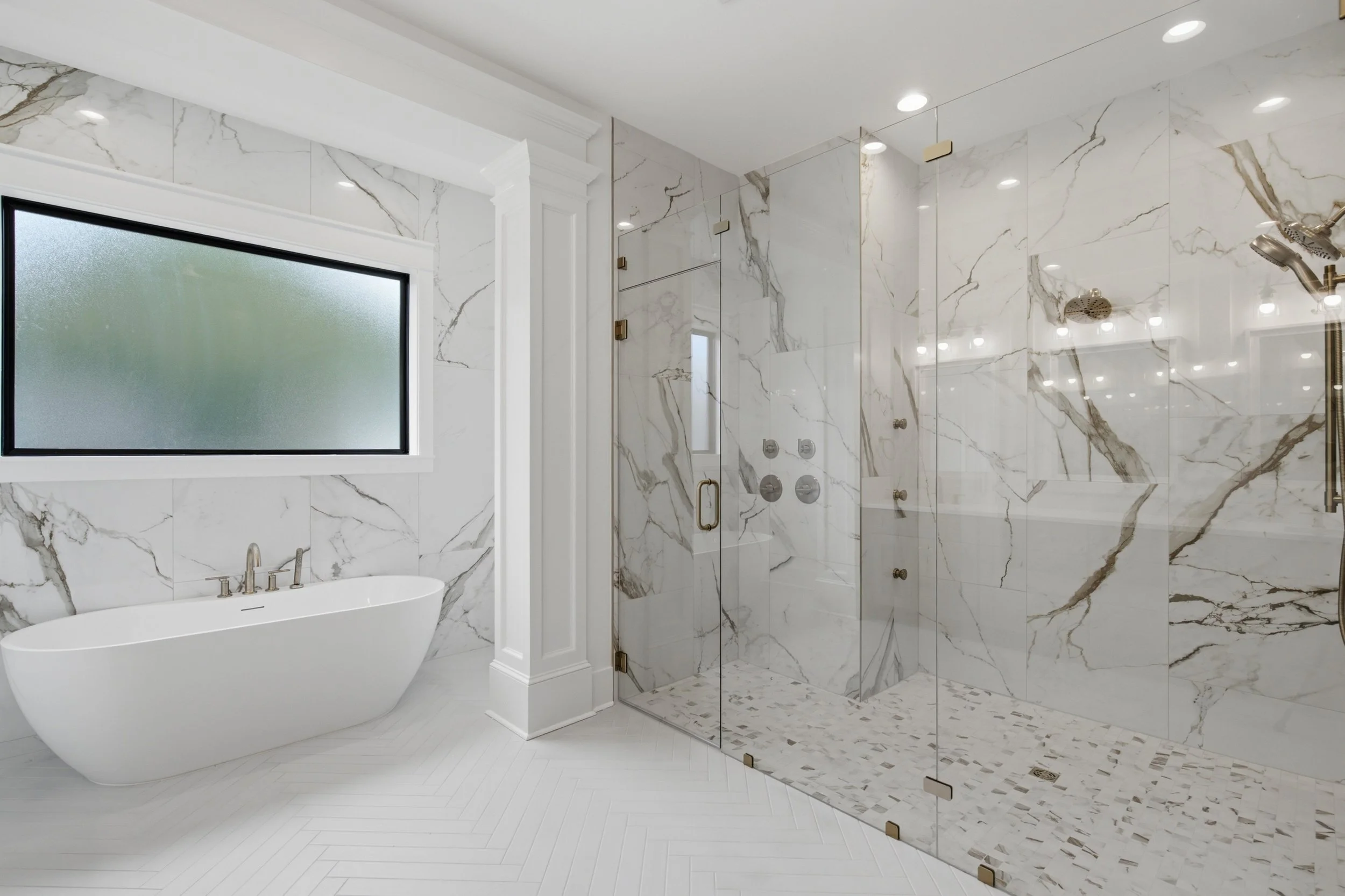 A luxurious bathroom with a freestanding bathtub, white marble walls with dark veins, a large frosted window, and a spacious glass-enclosed shower with multiple showerheads and controls.