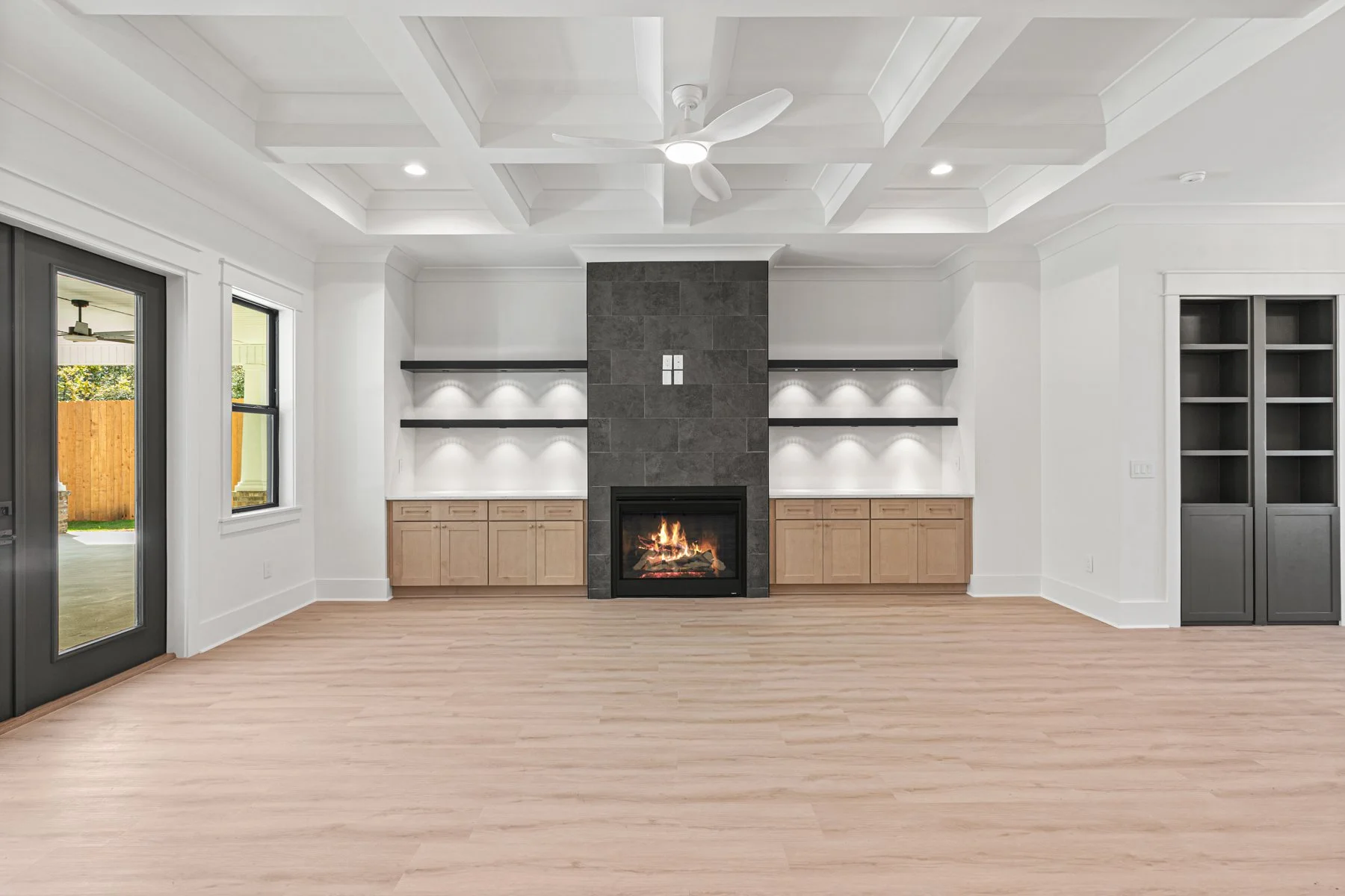 Living room with white walls and ceiling, featuring a grey fireplace with a fire burning, flanked by wooden cabinets and floating shelves with built-in lights. A ceiling fan with white blades is installed, and there are large windows and a glass door leading outside.