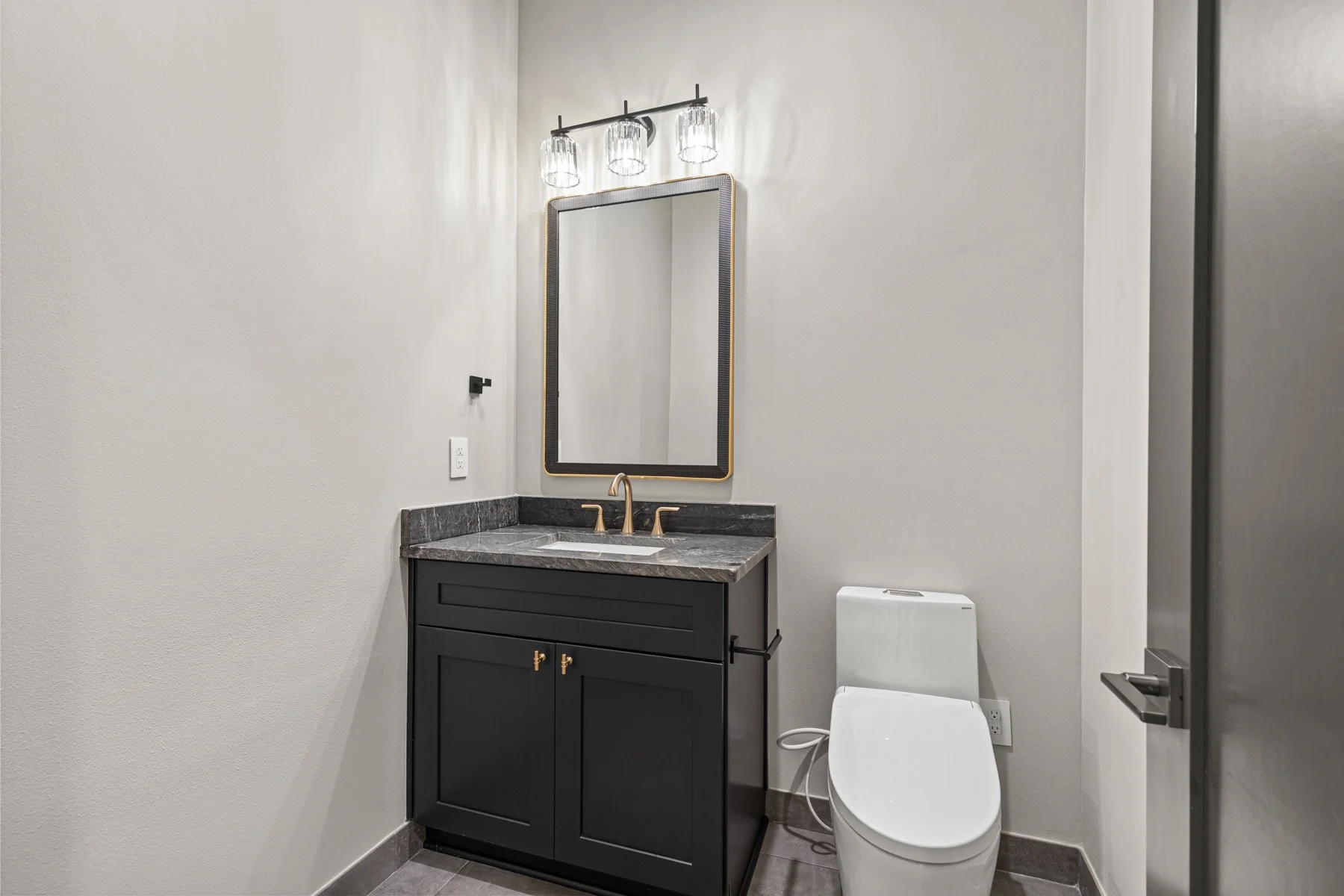 Modern bathroom with dark vanity, marble countertop, gold fixtures, large mirror, gray walls, toilet, and overhead light fixture.