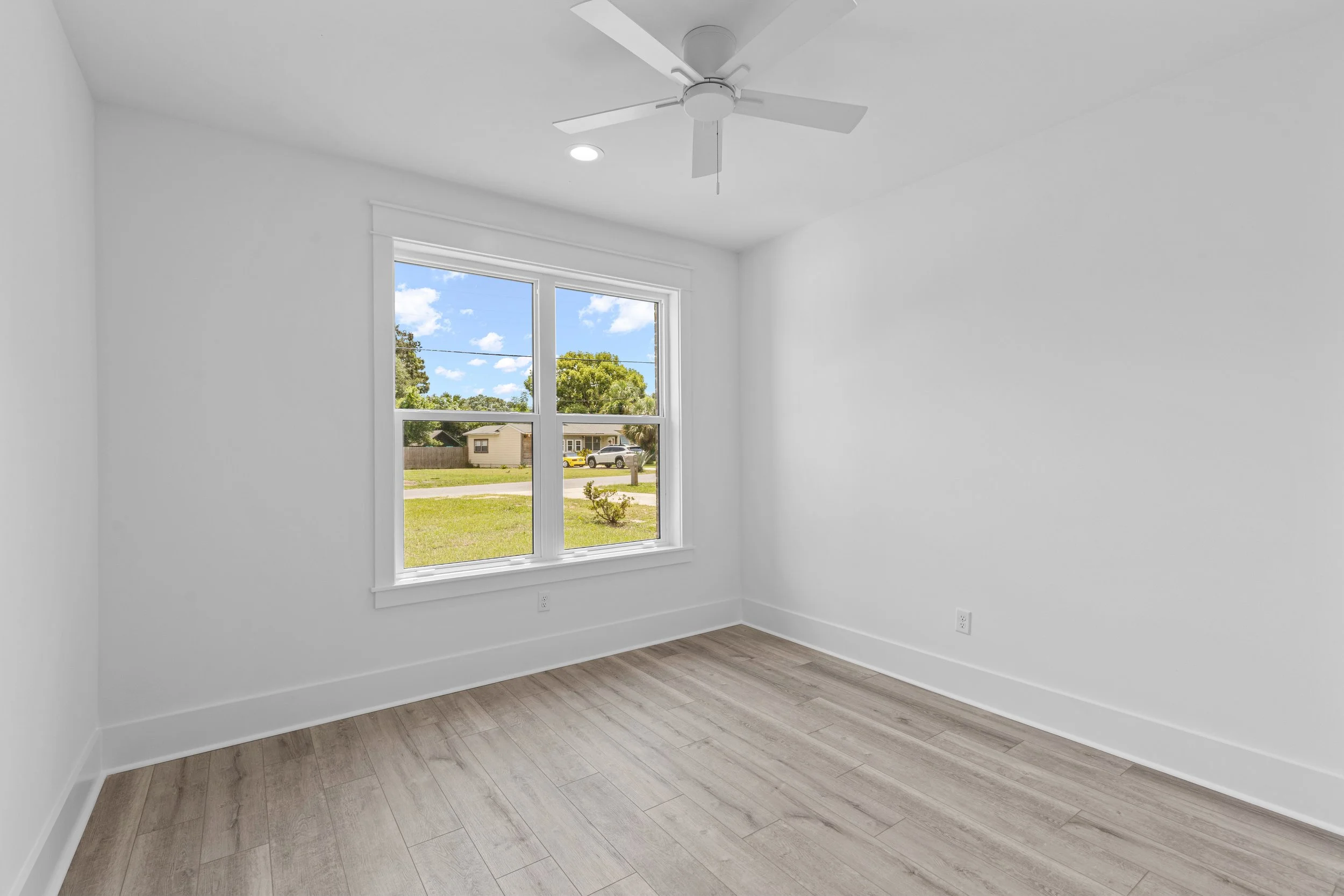 Empty room with white walls, a large window showing a sunny outdoor scene with trees, another house, and a street. The room has light wood flooring and a ceiling fan.