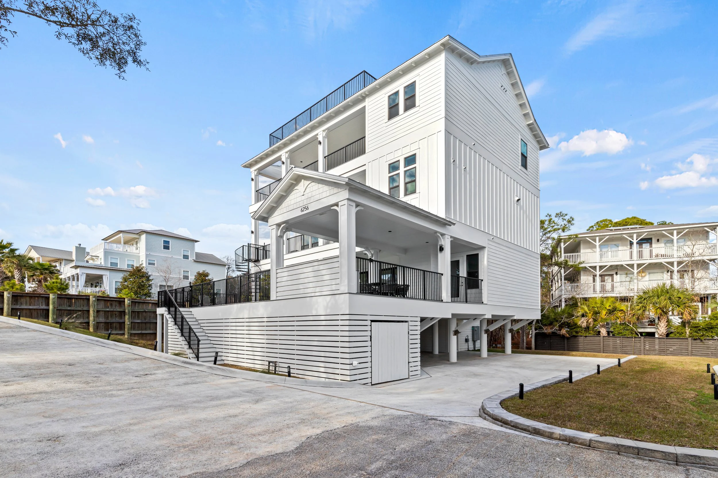 A white multi-story house with a modern design and black railings, elevated on stilts, with a parking area underneath, and other similar houses in the background under a blue sky with clouds.