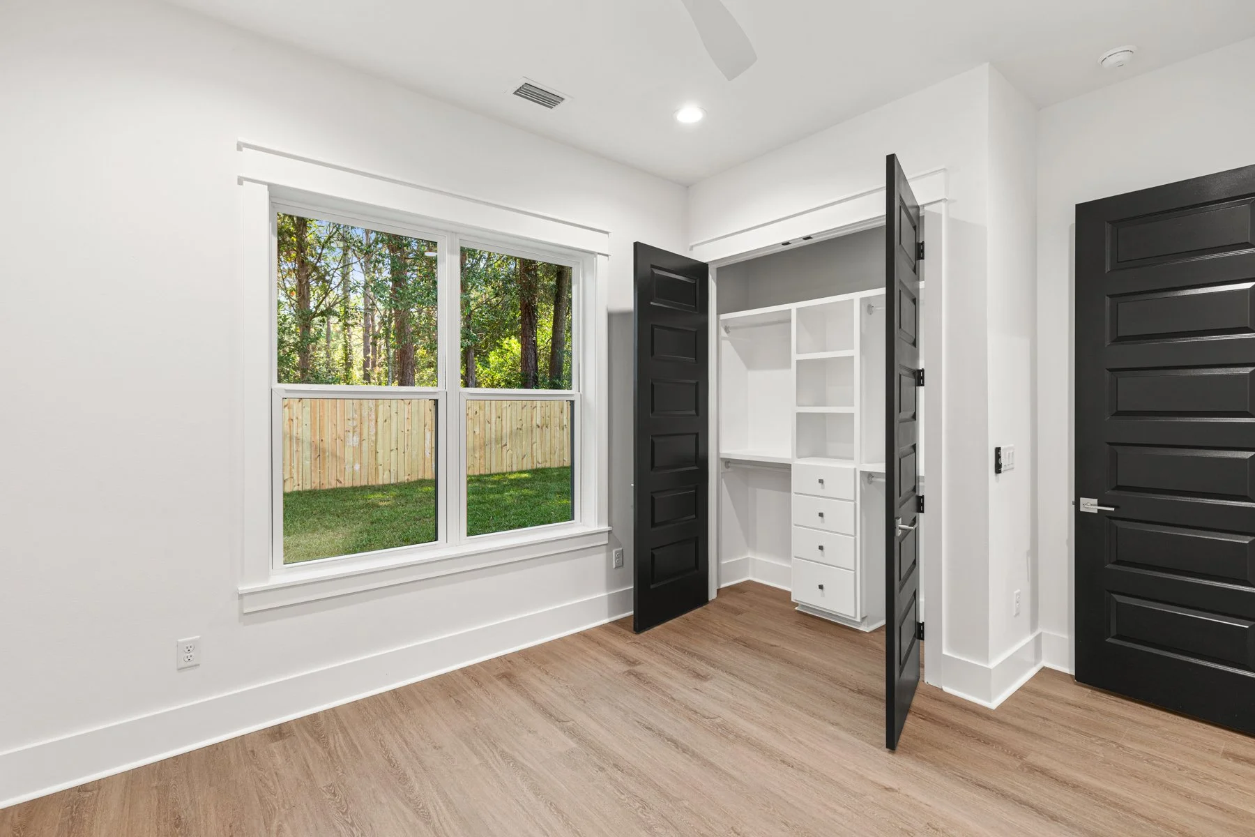 Empty room with white walls, a window looking out to a backyard with trees and a wooden fence, a built-in white closet with black interior doors, and wood flooring.