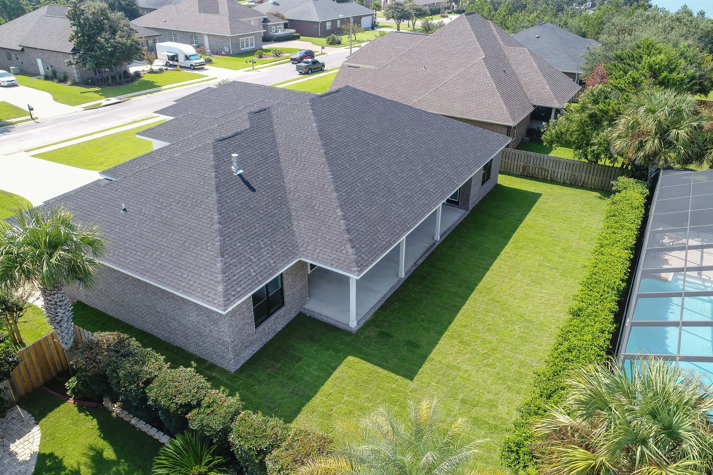 Aerial view of a suburban neighborhood with multiple houses, well-maintained lawns, trees, fences, and a backyard with a patio and pool.