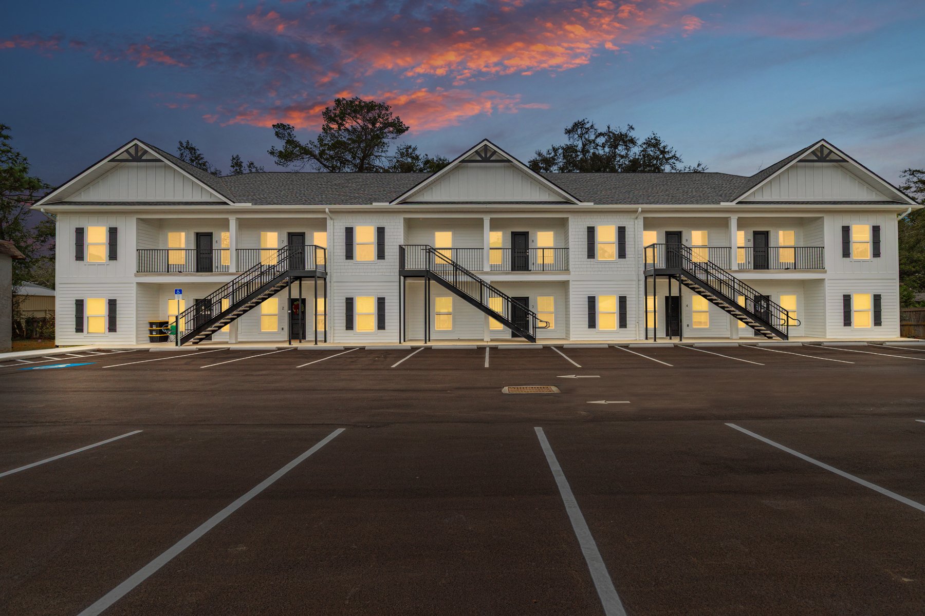 A two-story apartment complex at dusk with lit windows, exterior staircases, and an empty parking lot in front, under a dotted sky with pink and purple clouds and trees in the background.